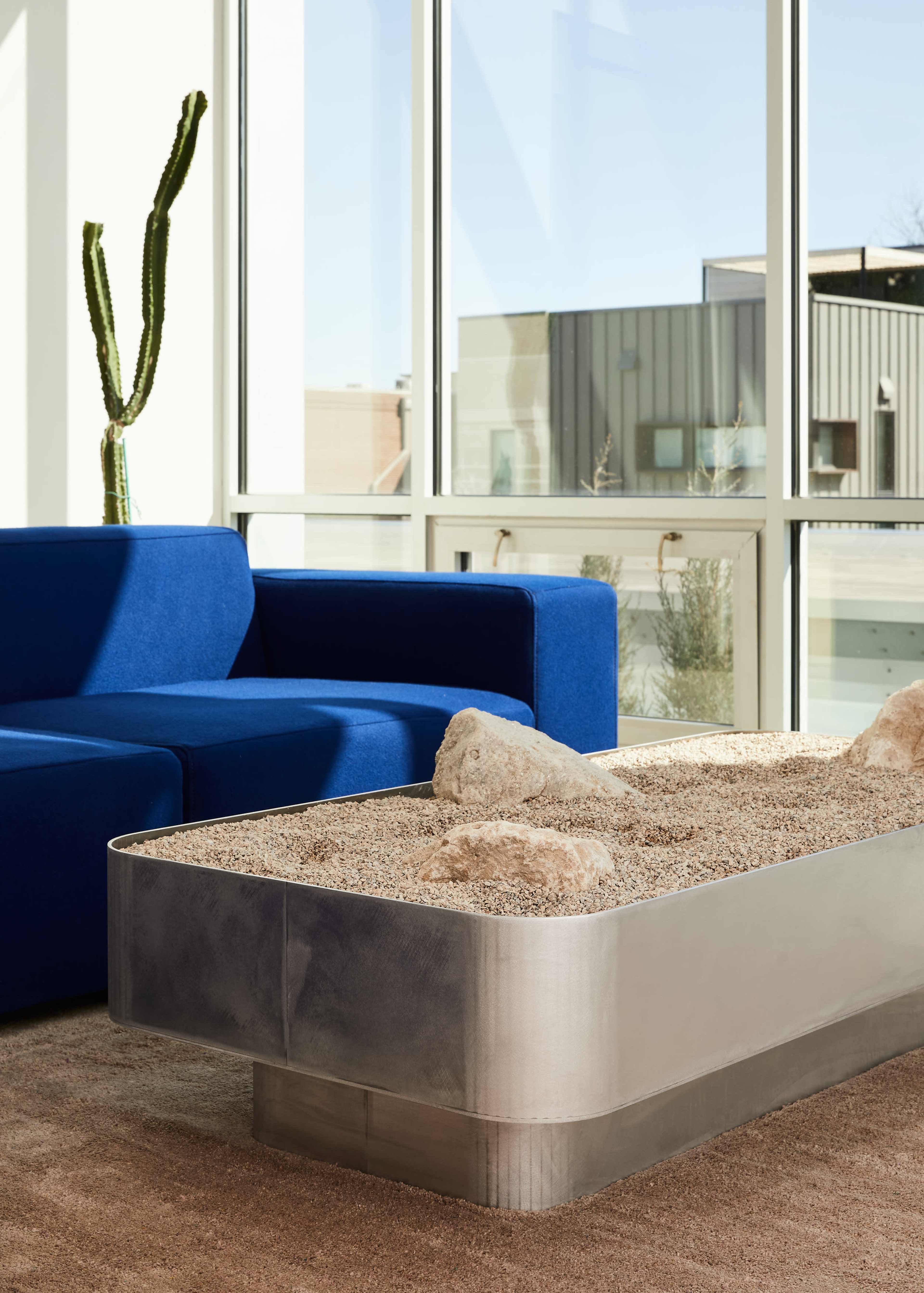 A blue couch is positioned next to a rectangular coffee table filled with sand and rocks, set in a room with large windows and a view of a modern building.
