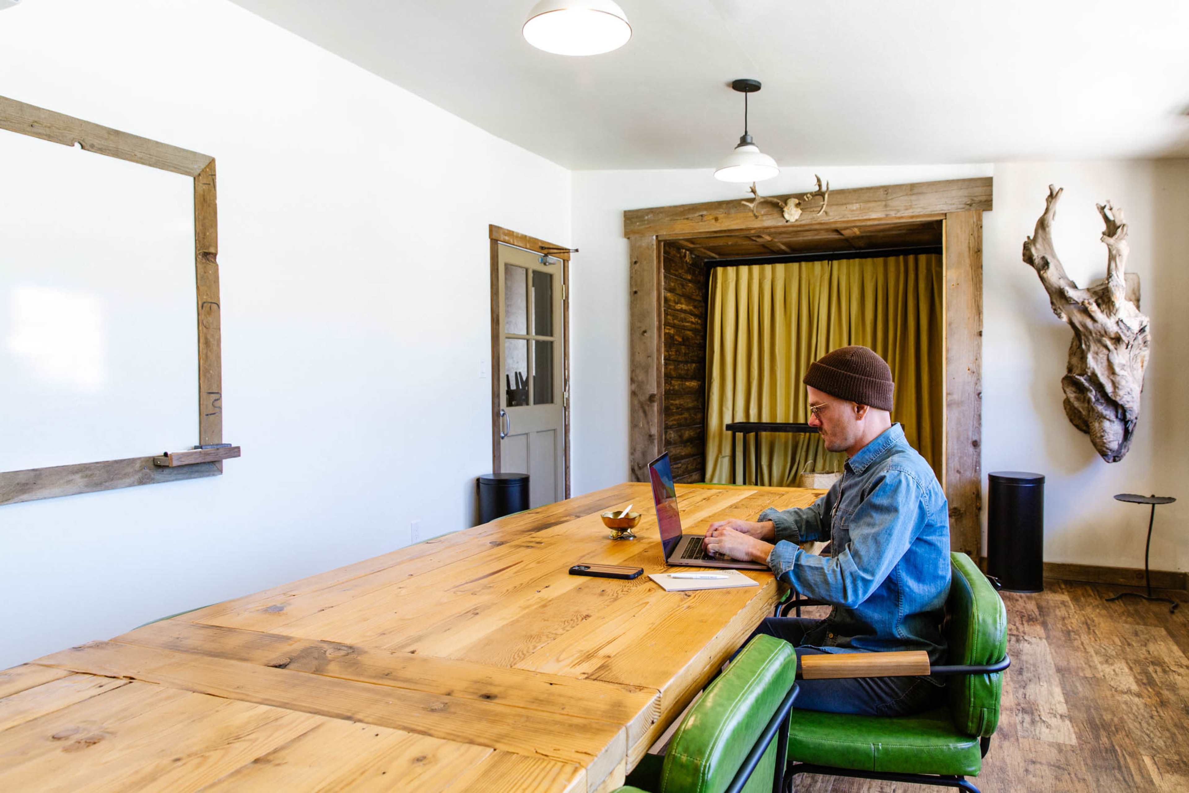 A person works on a laptop at a large wooden table in a bright, minimalist room with wooden accents and a curtain in the background.
