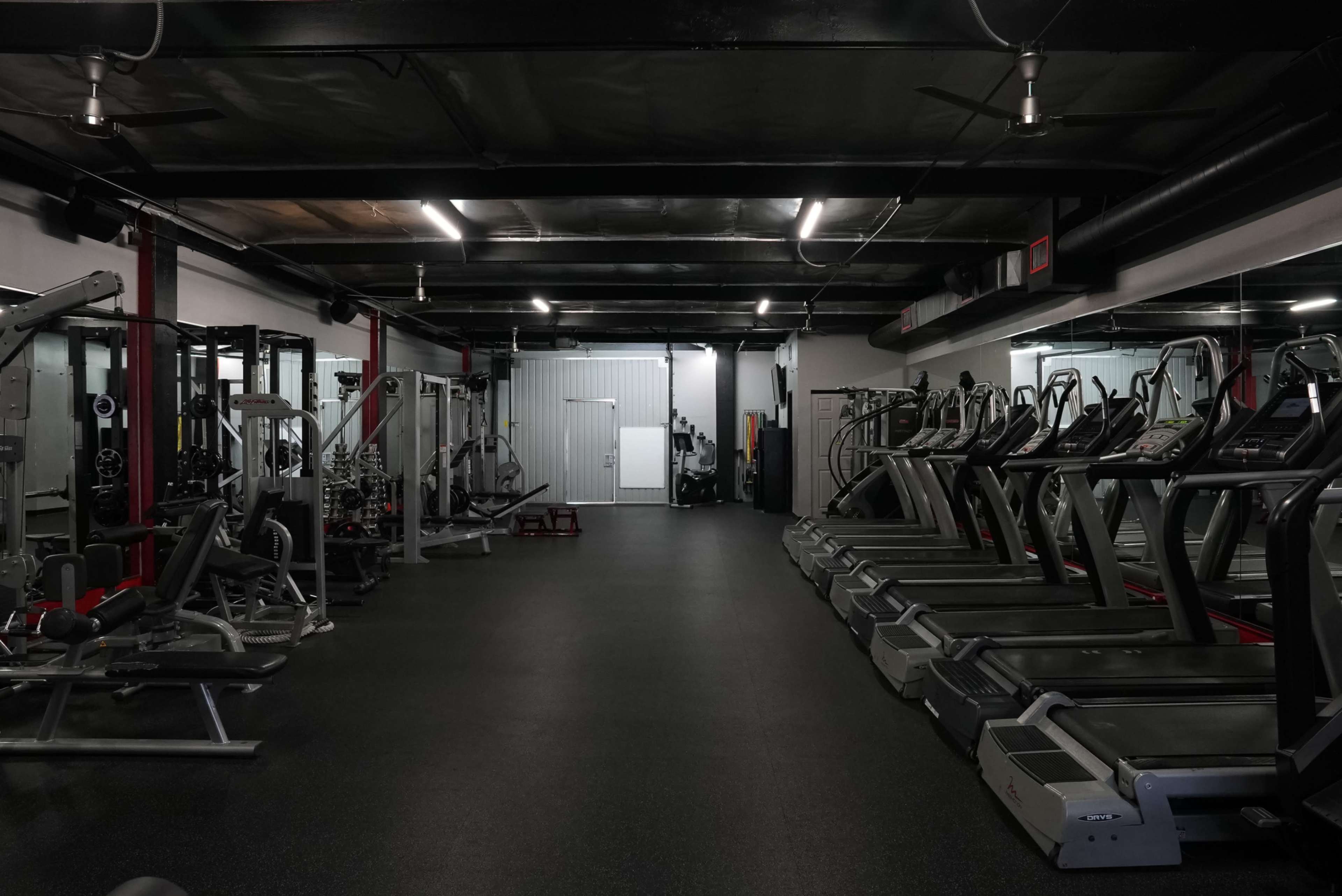 An empty gym with various exercise equipment, including treadmills and weight machines, arranged along the walls under artificial lighting.