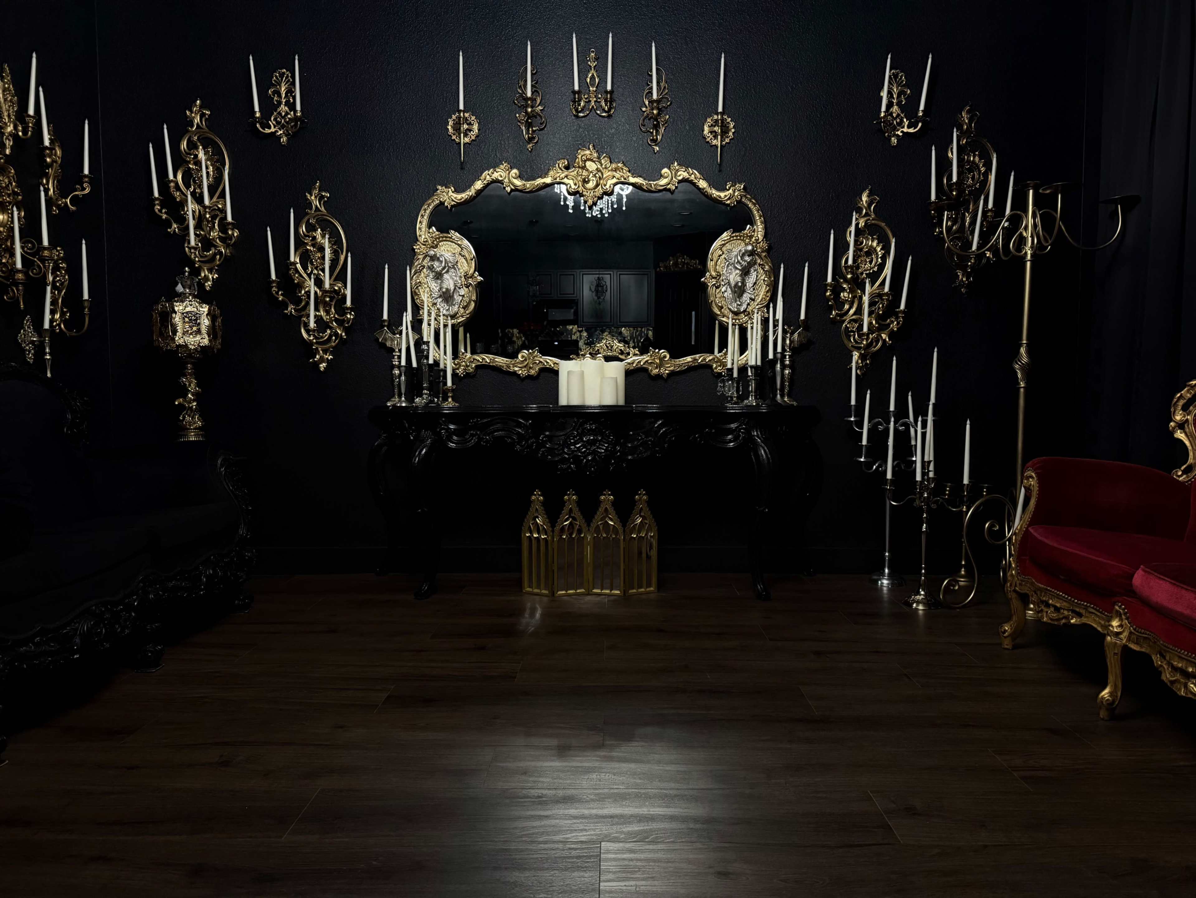 The room features ornate gold wall sconces, a large gilded mirror, and a black table adorned with candles, surrounded by dark furniture in a dimly lit setting.