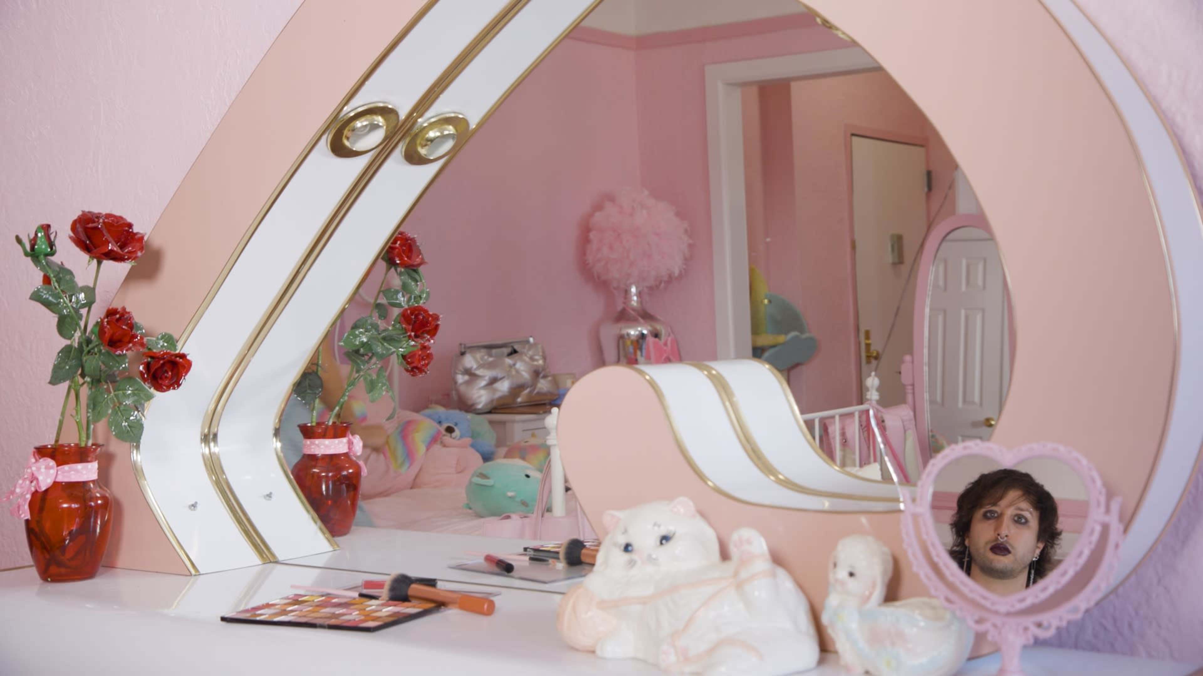 A vanity mirror with a pink and white design reflects a softly lit room that features stuffed animals, floral decorations, and cosmetics.