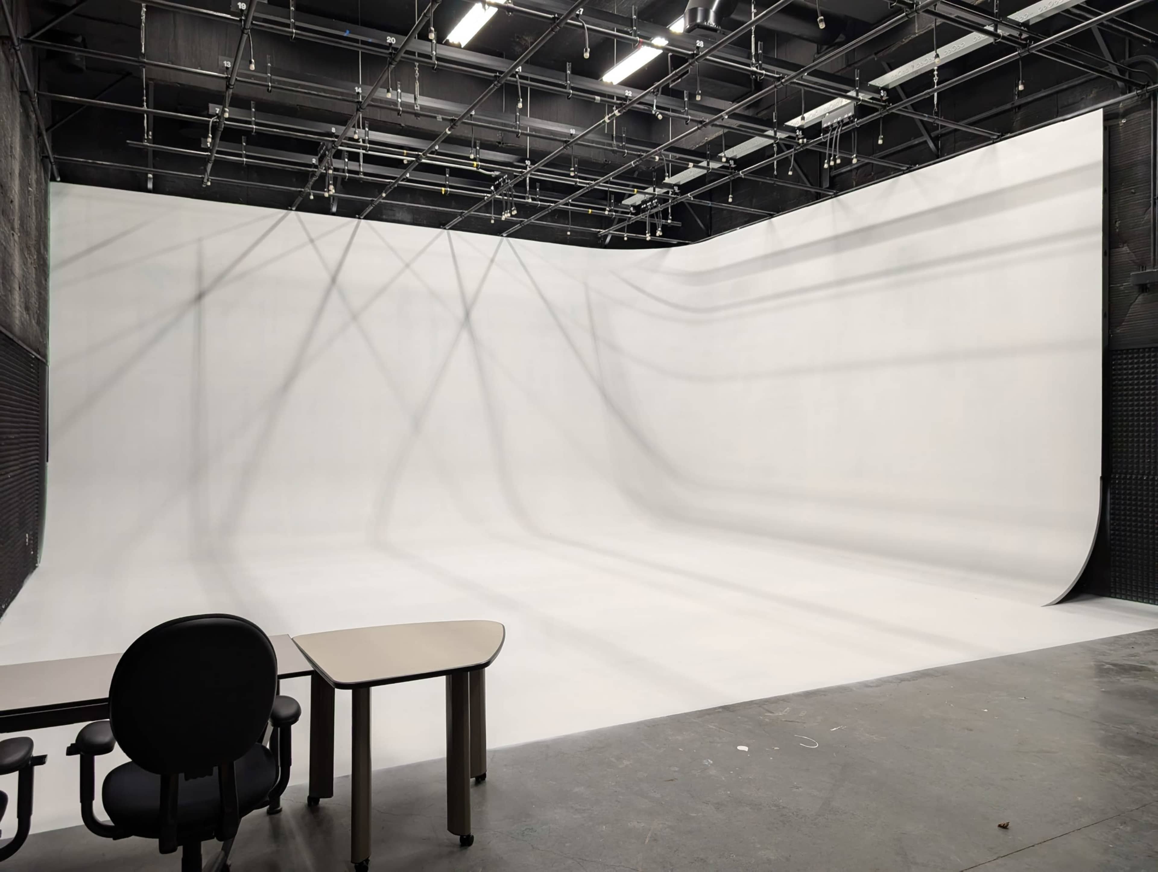 The image shows an empty studio space with a seamless white backdrop and a table with a chair on one side.