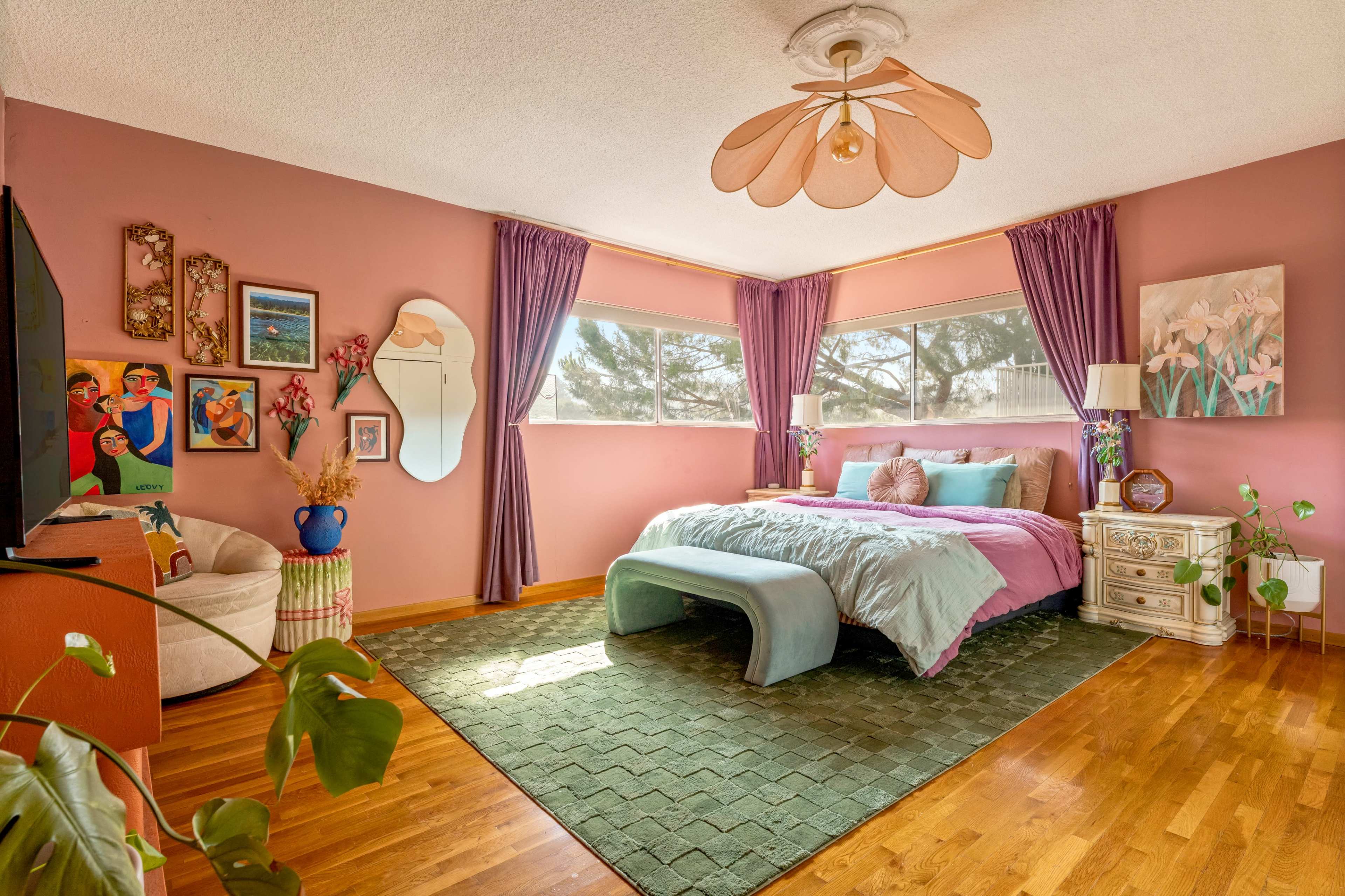 The image shows a colorful bedroom with pink walls, large windows, a bed with a light purple comforter, and a green area rug on a wooden floor.