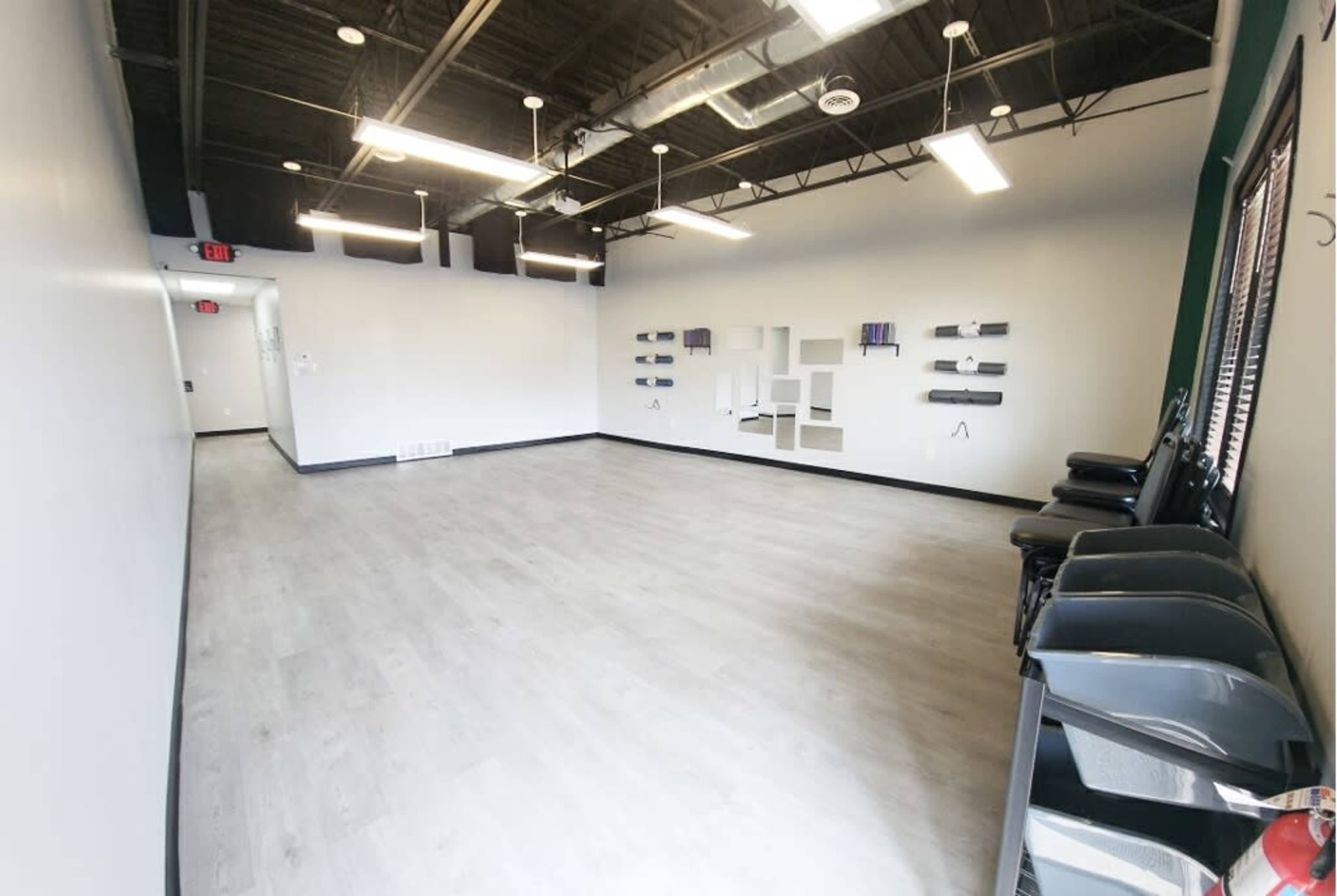 A spacious, empty room with light-colored flooring, exposed walls, and several chairs aligned against one side.