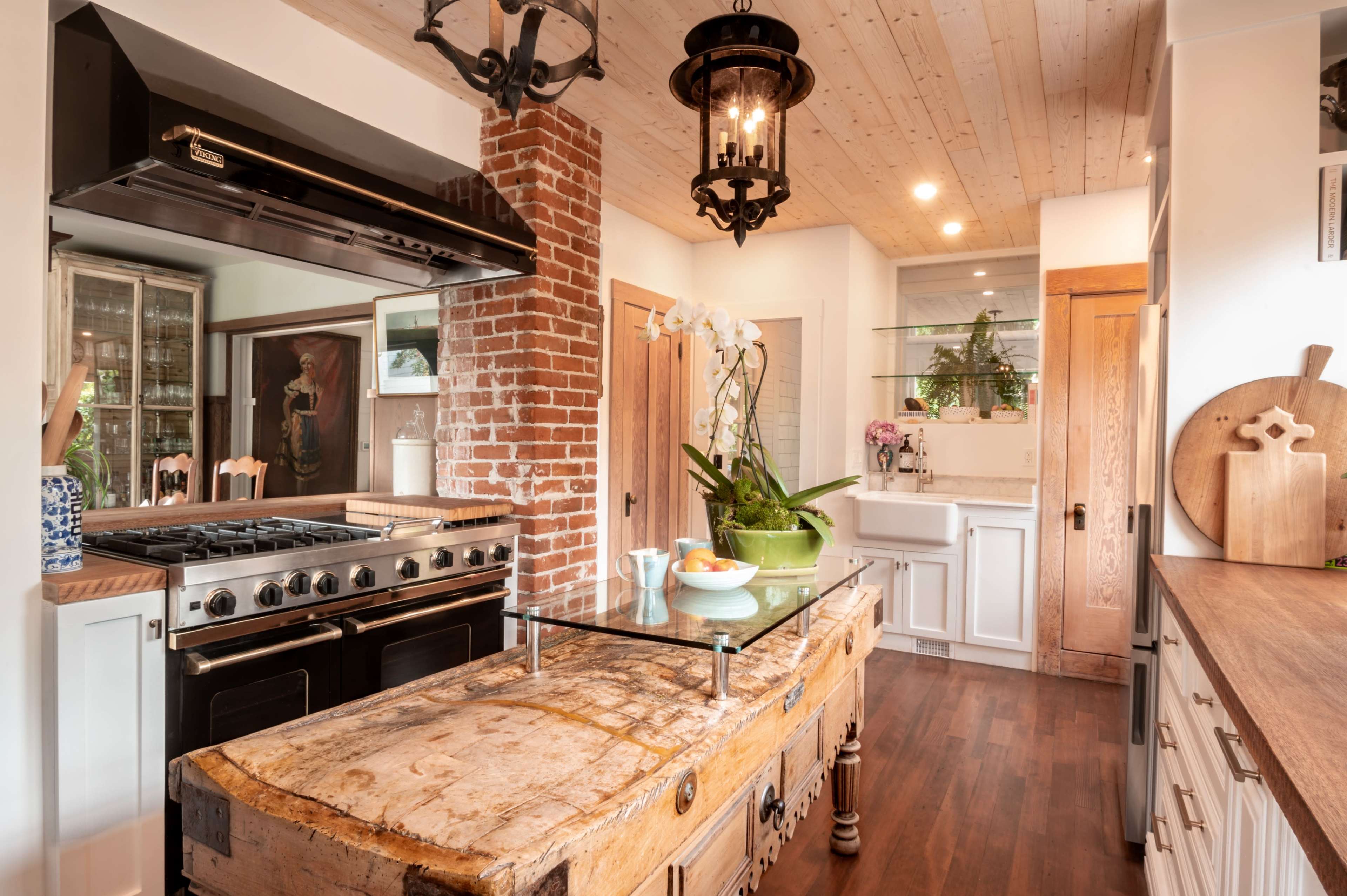 The kitchen features a large central island made of wood, brick walls, and modern appliances.