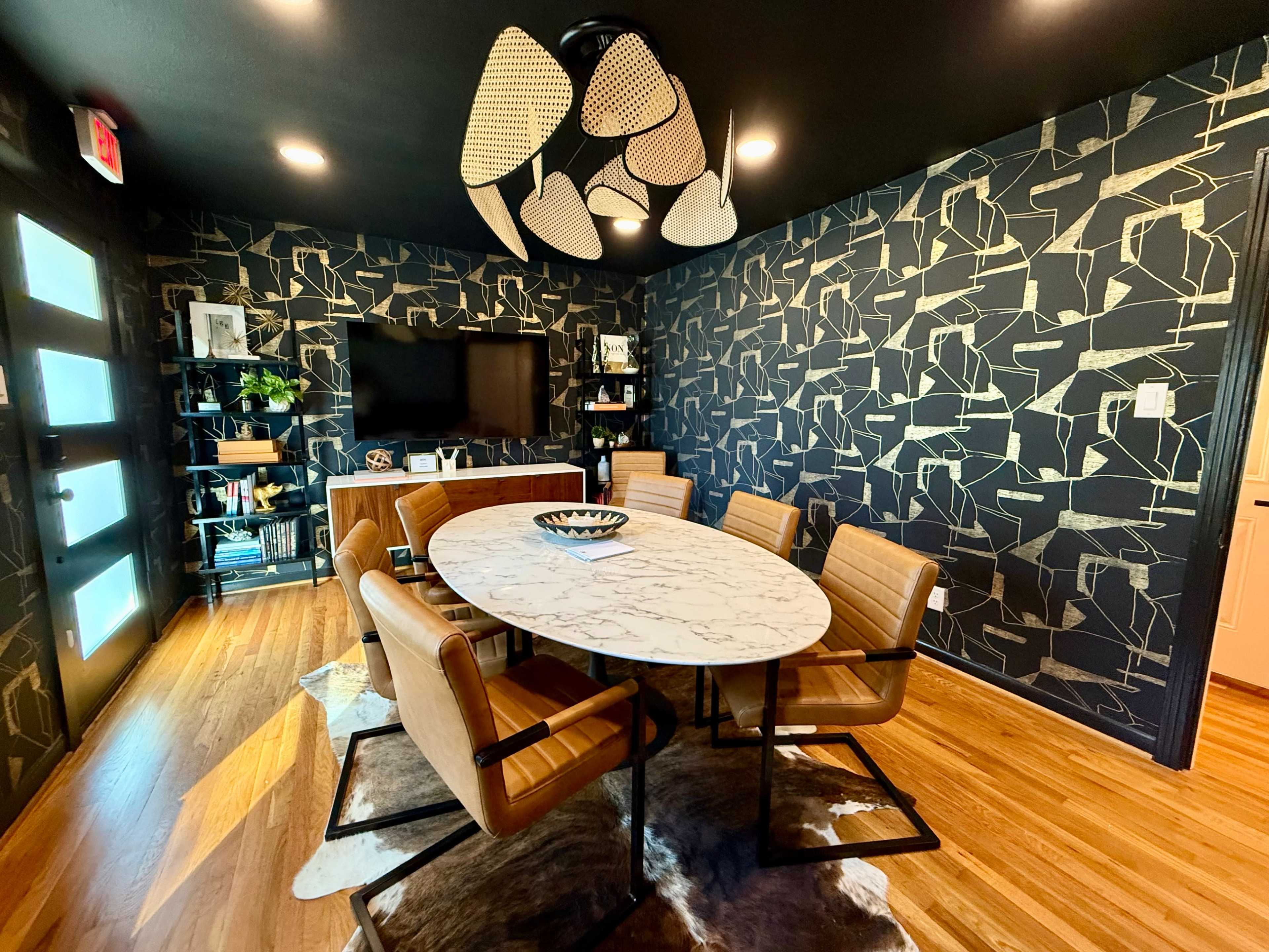 A modern dining room features a round marble table surrounded by four tan chairs, with a patterned black wall, a TV on one side, and a decorative light fixture above.