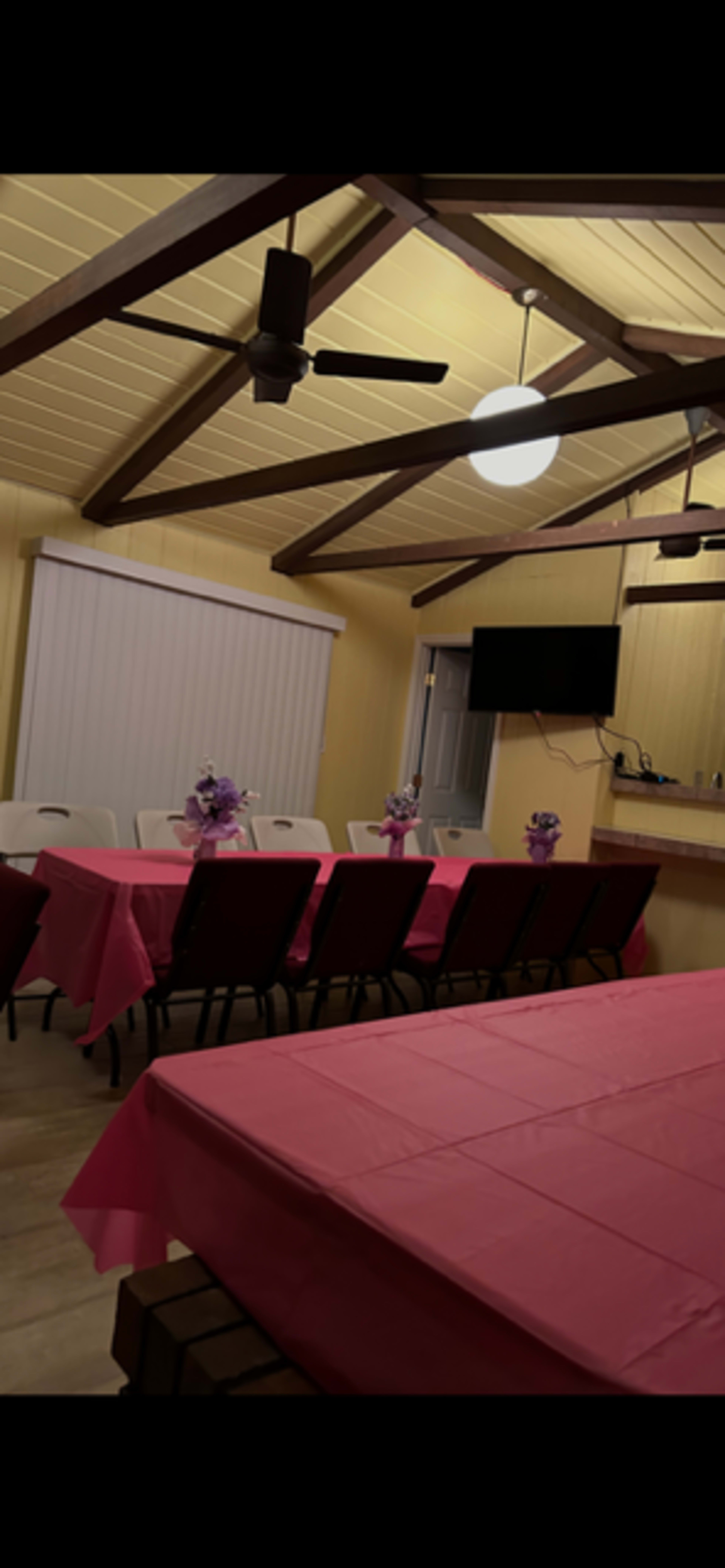 The image shows a dining area with several tables covered in pink tablecloths, decorated with flower centerpieces, and a television mounted on the wall.