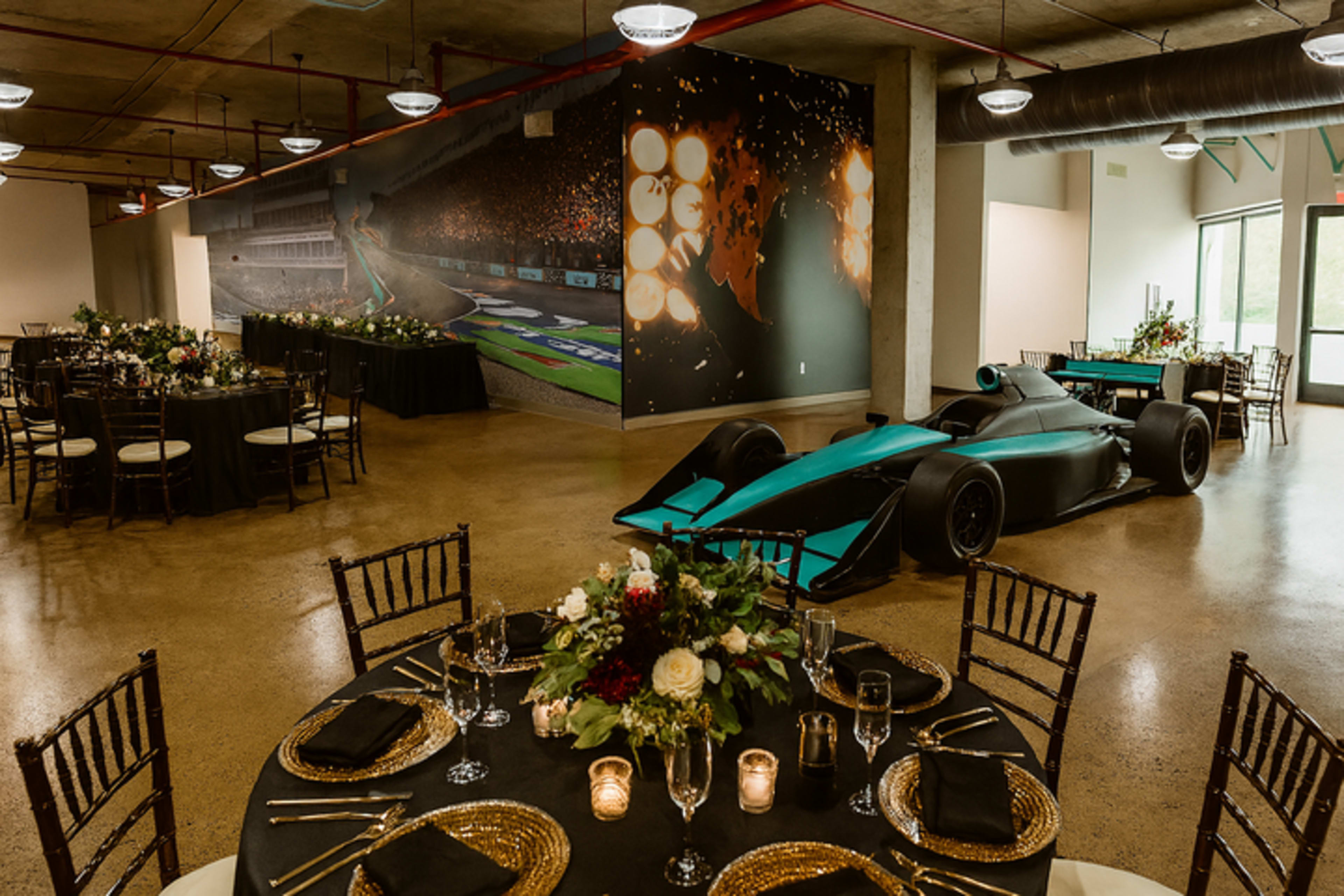 The image features a modern event space with elegantly arranged tables, adorned with floral centerpieces and a large racing car displayed prominently against a mural of a racetrack.