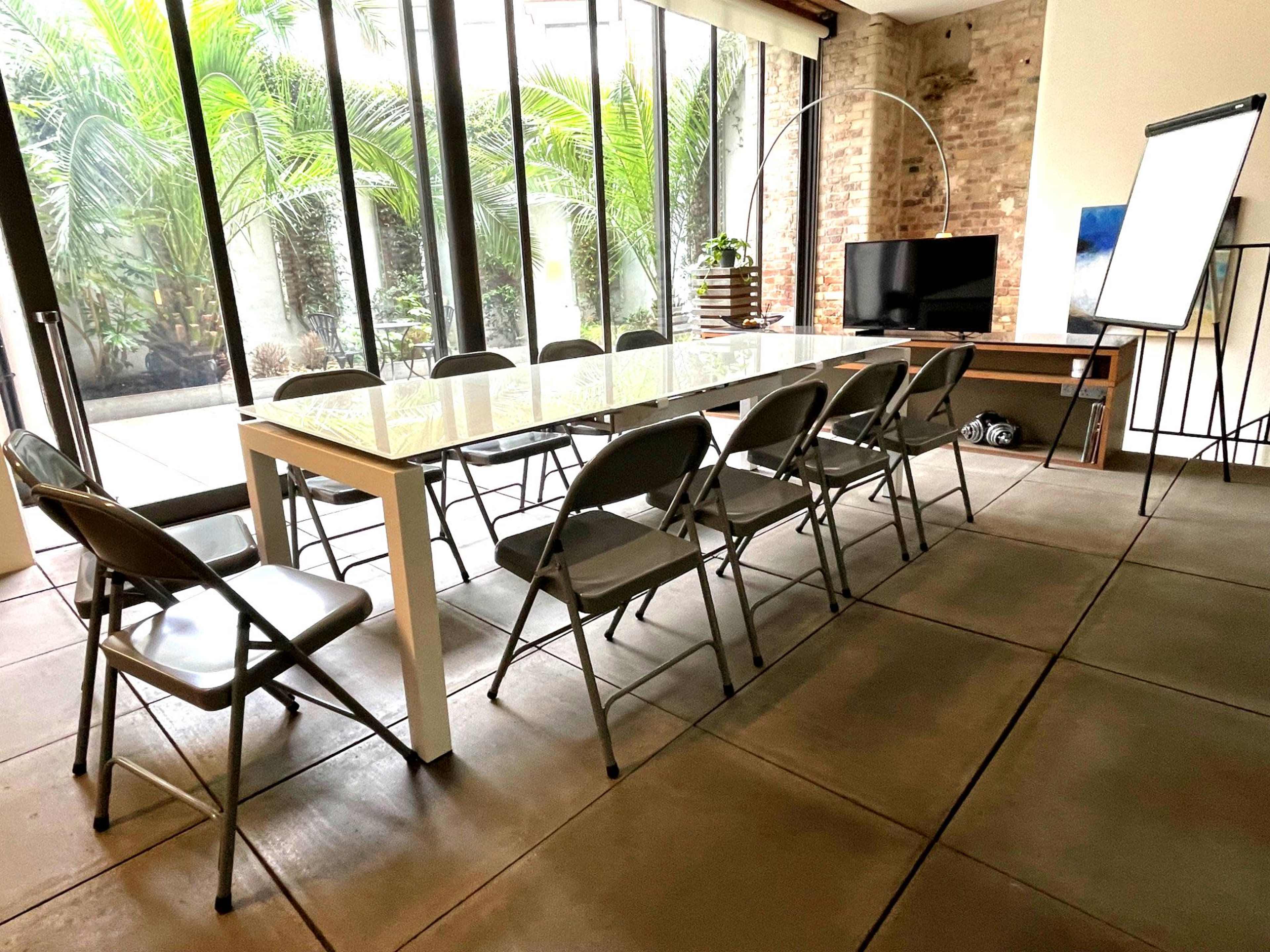 A long white table with eight folding chairs is set up in a bright room featuring large glass windows and greenery outside.