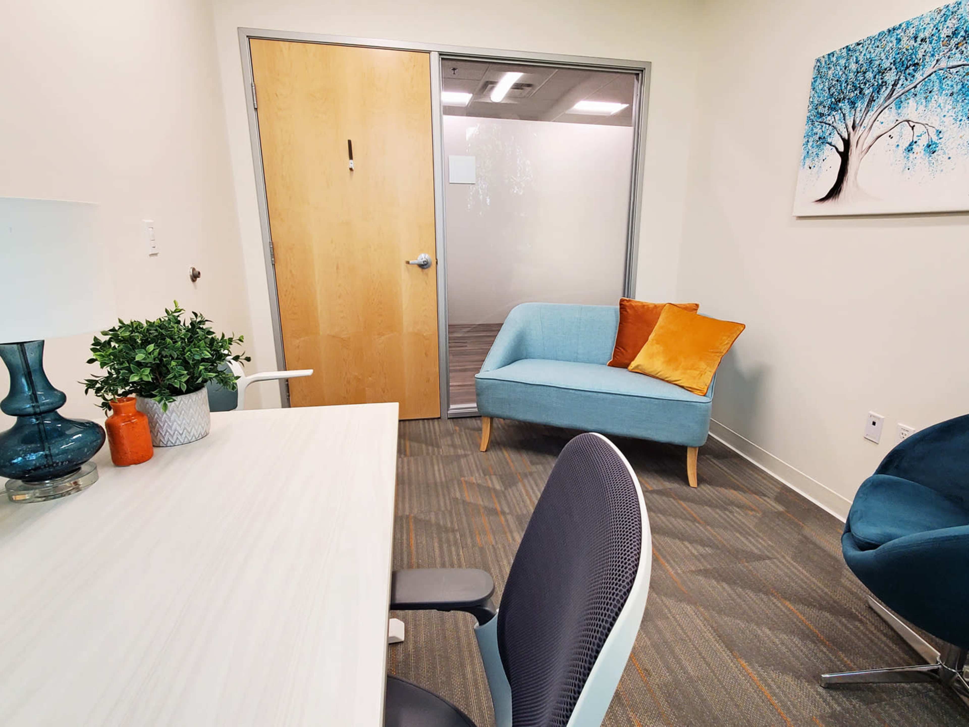 A modern office space with a desk and chair, a small couch with a pillow, and a decorative painting on the wall.
