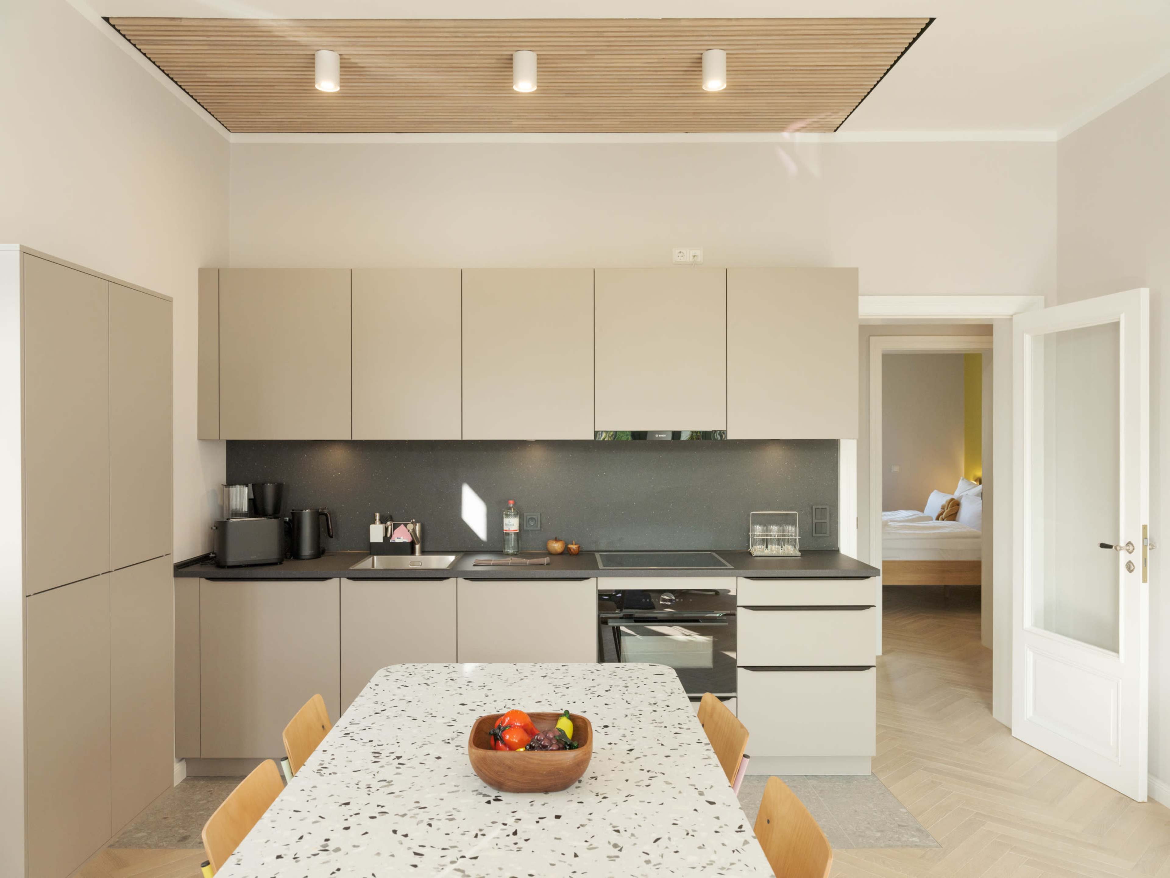 A modern kitchen features a light-colored cabinetry setup, a central dining table with a bowl of fruit, and a doorway leading to a bedroom.