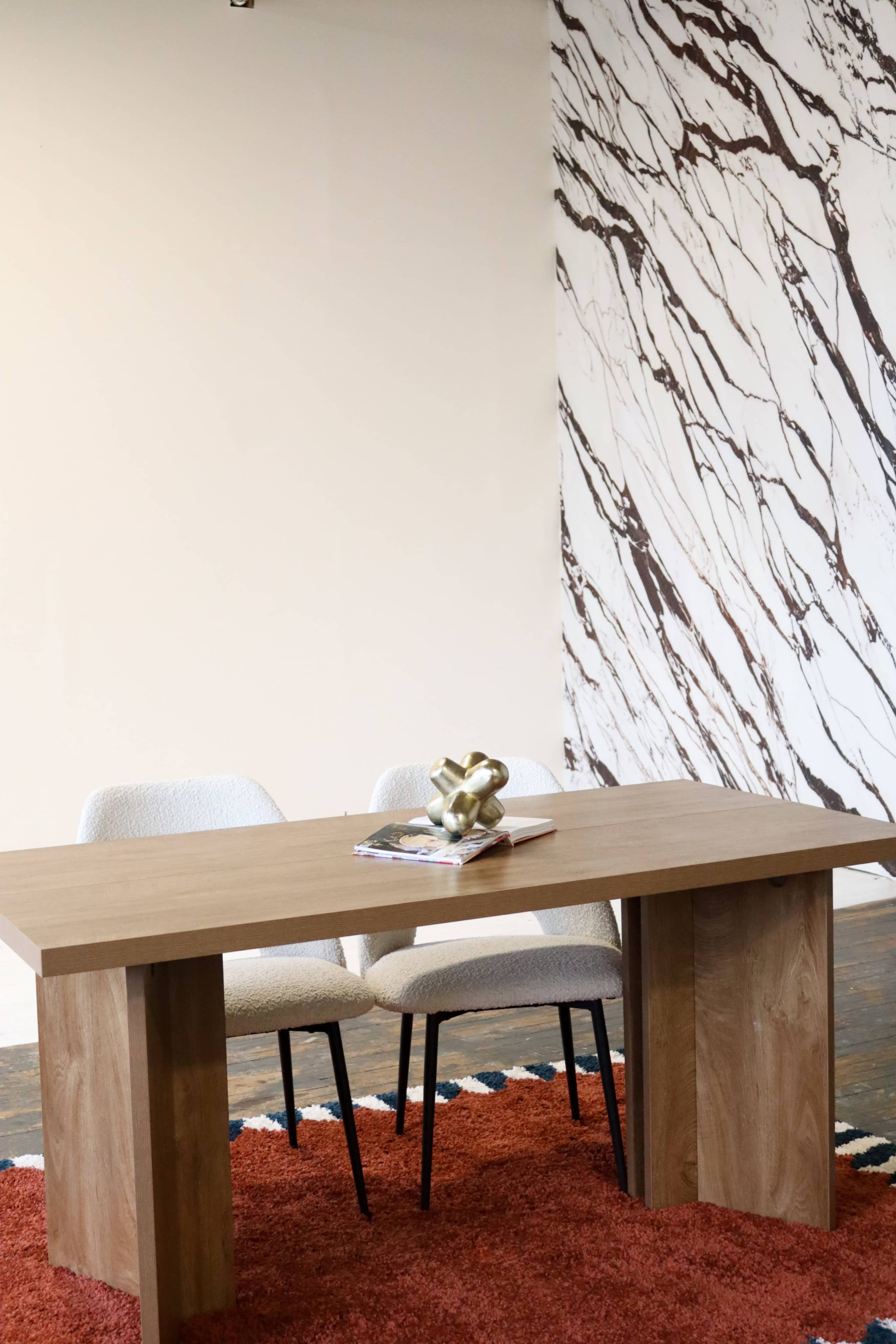 A wooden table with two chairs is set against a marbled wall and a textured rug.