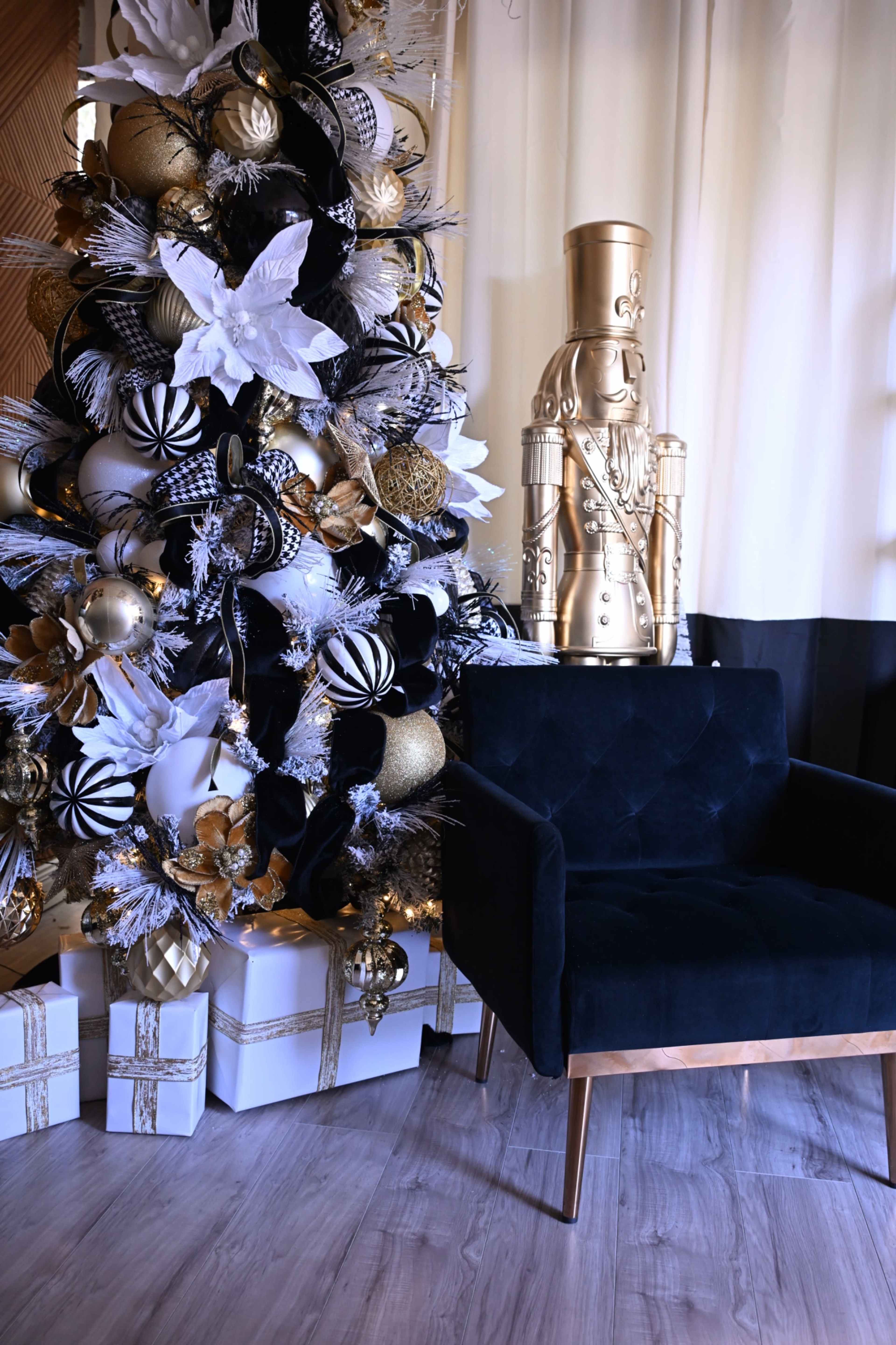 The image shows a decorated Christmas tree with black, gold, and white ornaments beside a black chair and a golden nutcracker figure against a light-colored backdrop.