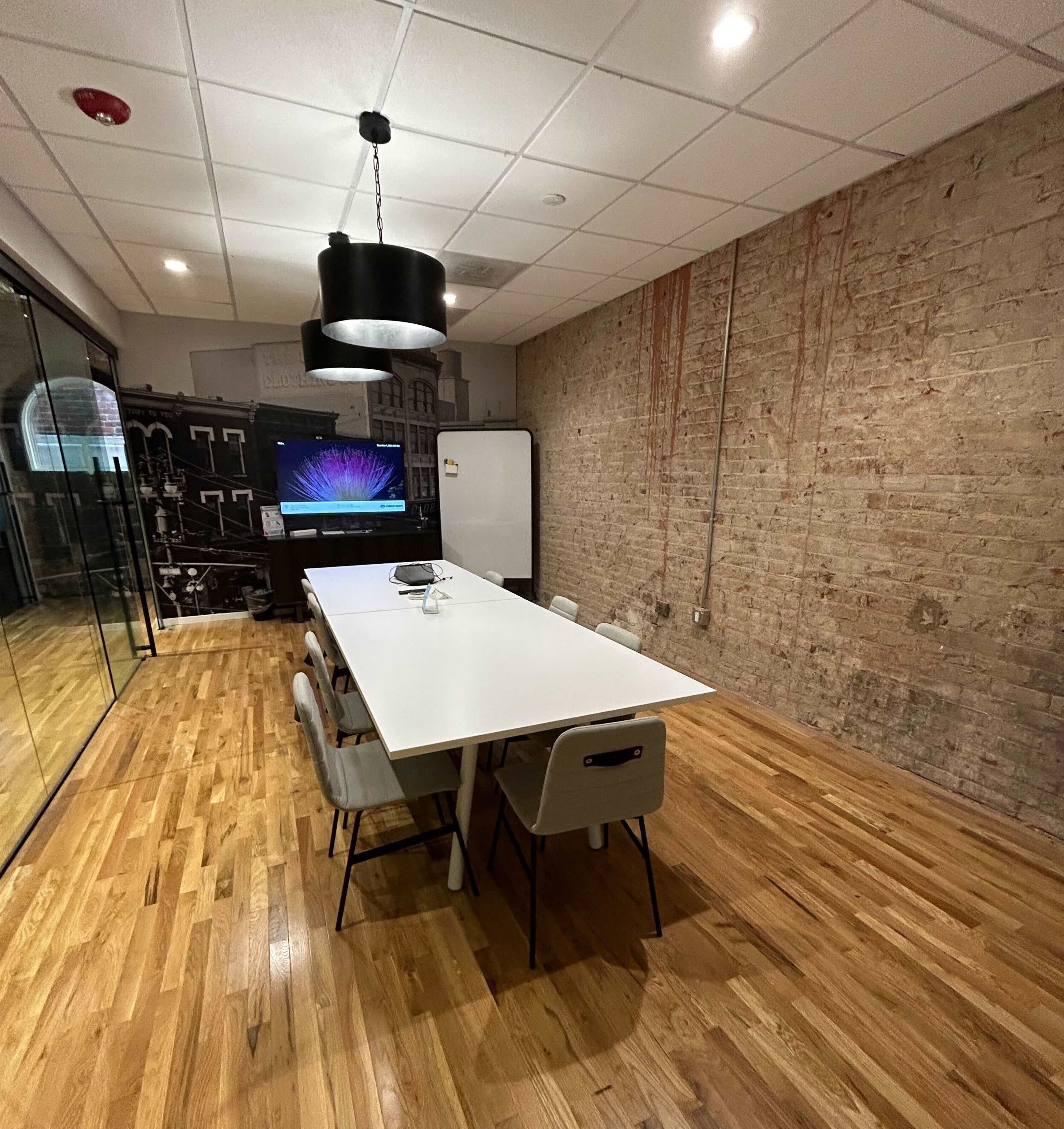 A modern conference room features a long white table surrounded by chairs, with a large screen on one wall and a brick accent wall.