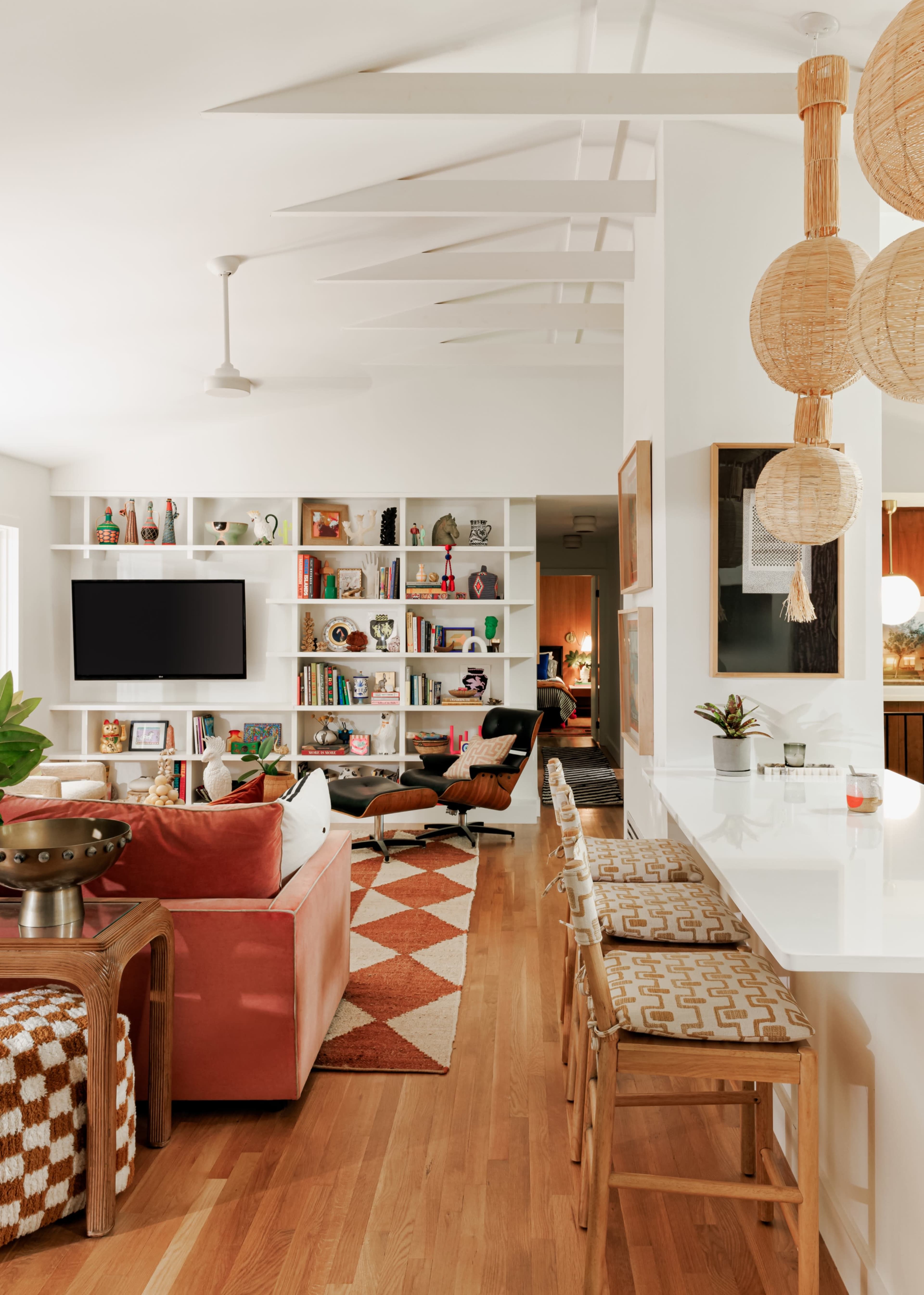 The image shows a modern living room with a pink sofa, a patterned area rug, a bookshelf filled with decorative items, and a dining area with bar stools.