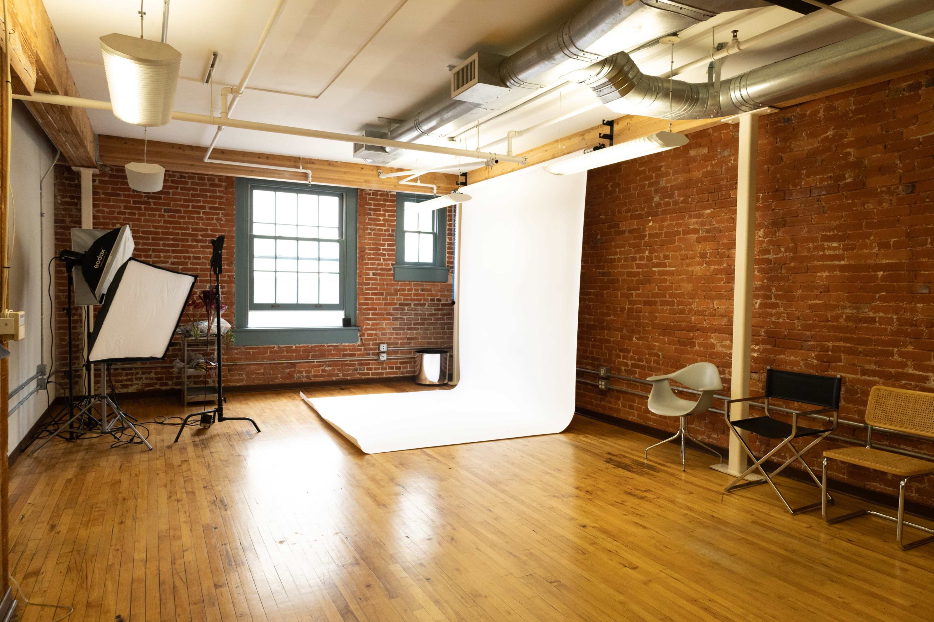 The image shows a spacious photography studio with exposed brick walls, wooden floors, and a white backdrop set up for shoots.