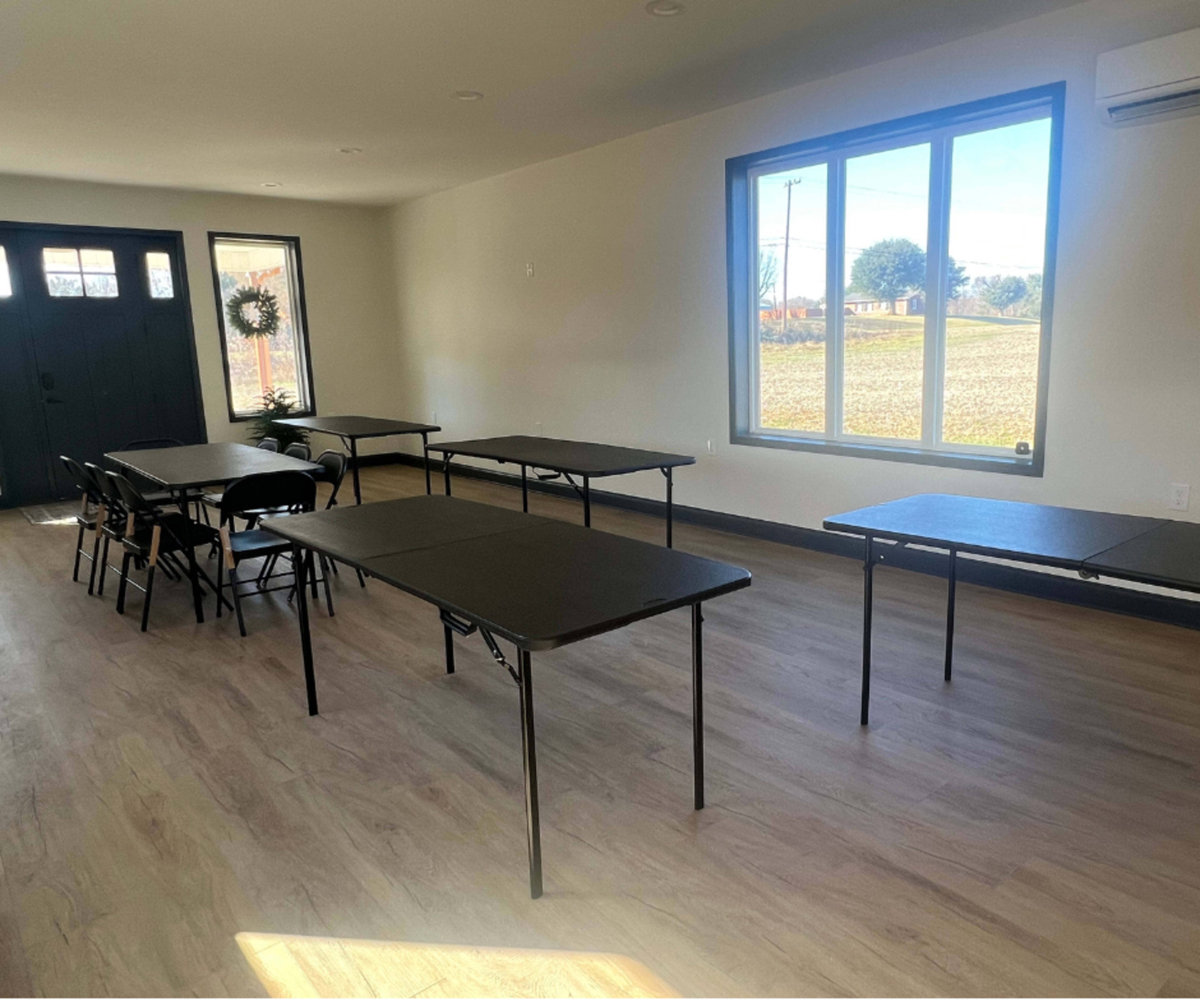 A room features several folding tables arranged in a spacious layout with large windows allowing natural light to enter.