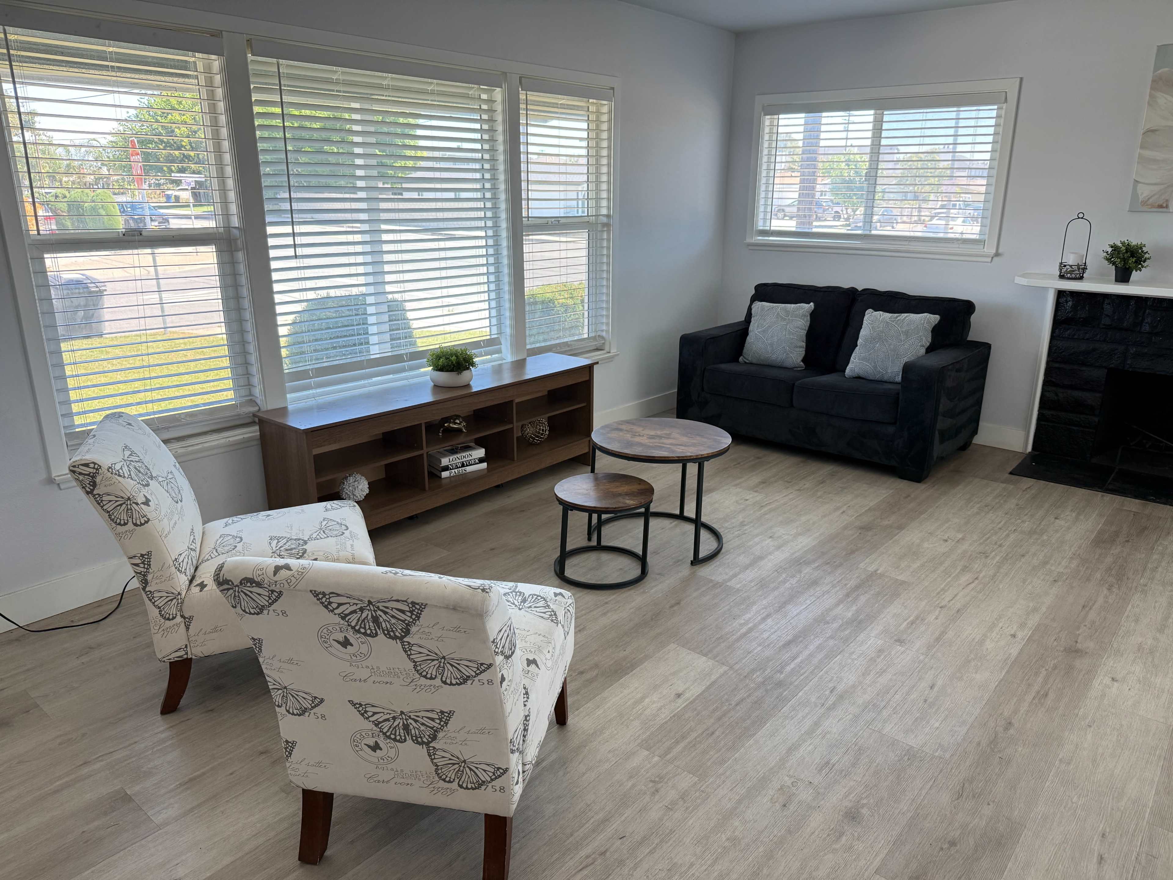 The image shows a living room with two patterned chairs, a black couch, and a coffee table, featuring large windows that let in natural light.