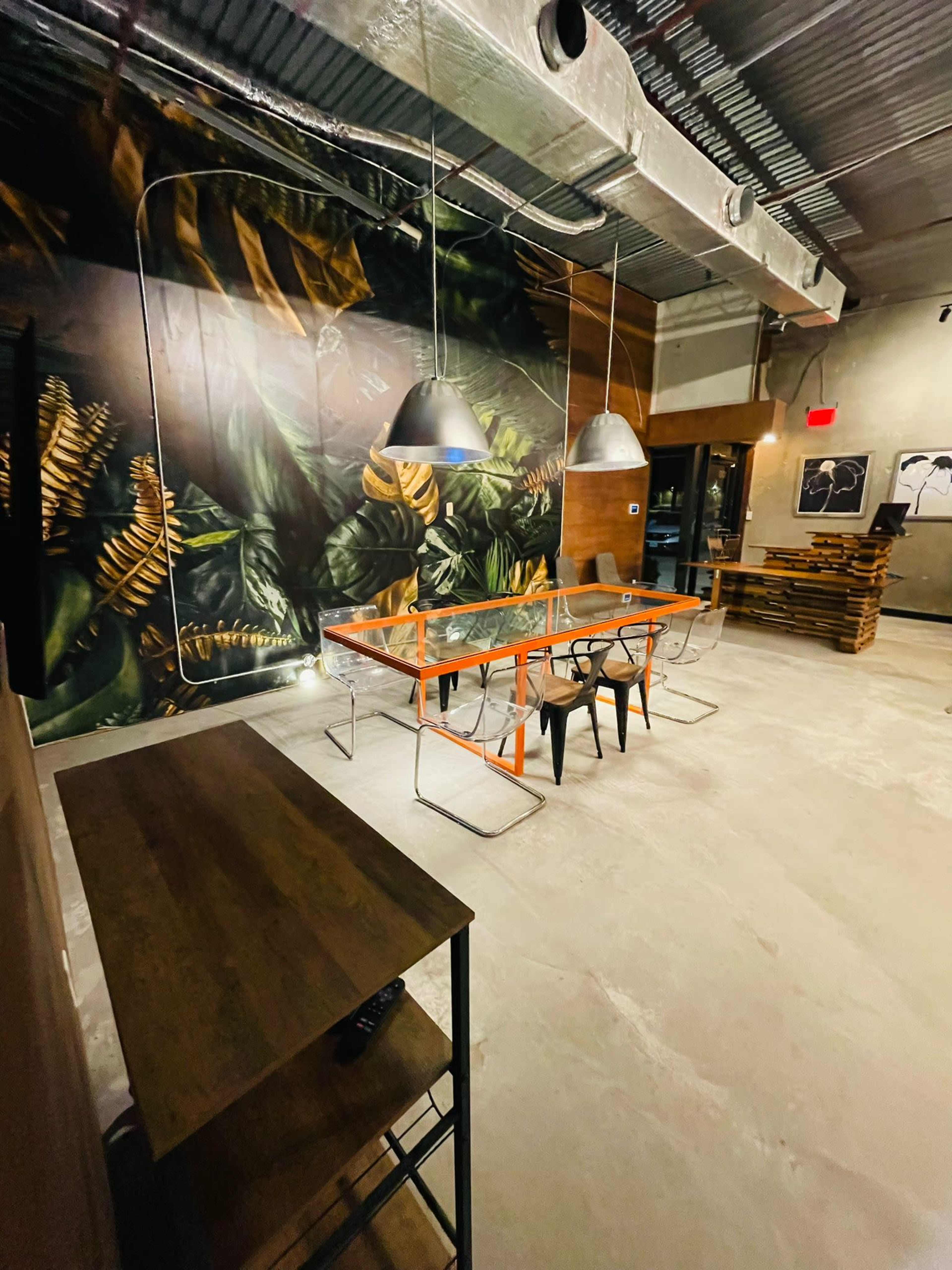 A modern interior space featuring a large table with an orange frame, black chairs, and a prominent mural of greenery on the wall.