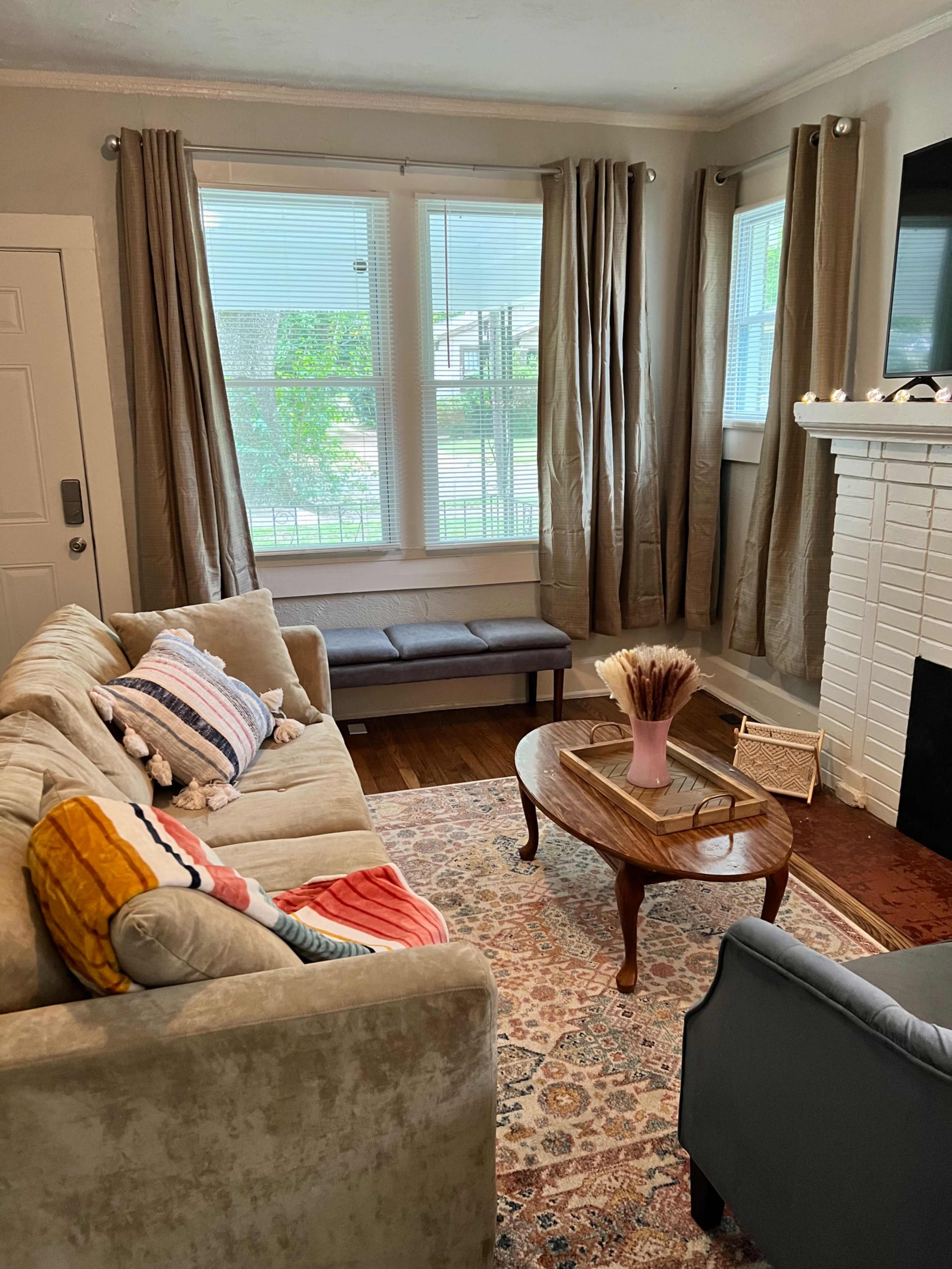 The image shows a living room with a beige sofa, a blue armchair, a wooden coffee table, and a white brick fireplace, all complemented by large windows draped with beige curtains.