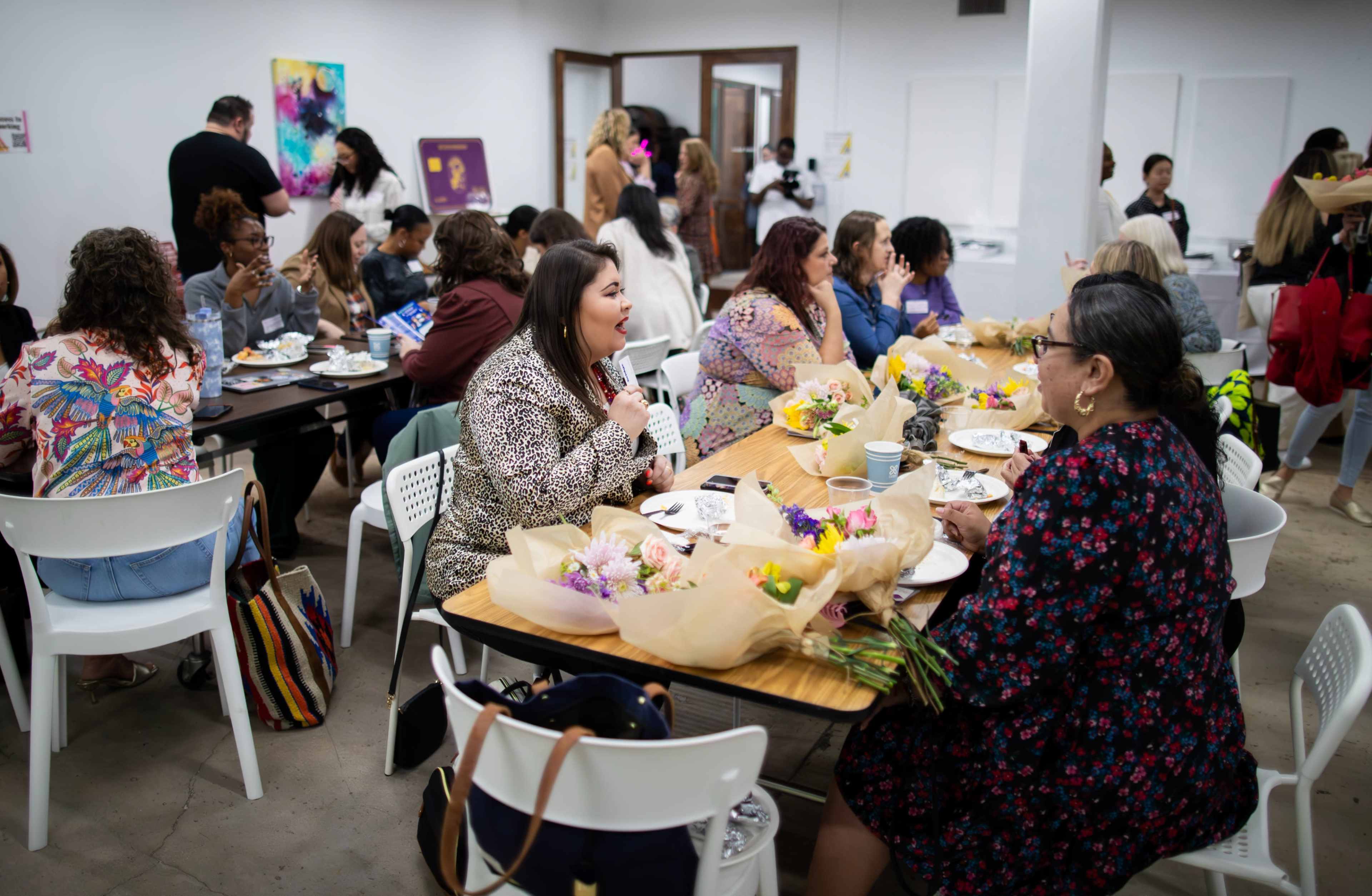 A group of people gathers around tables in a well-lit room, engaging in conversation while enjoying food and floral arrangements.