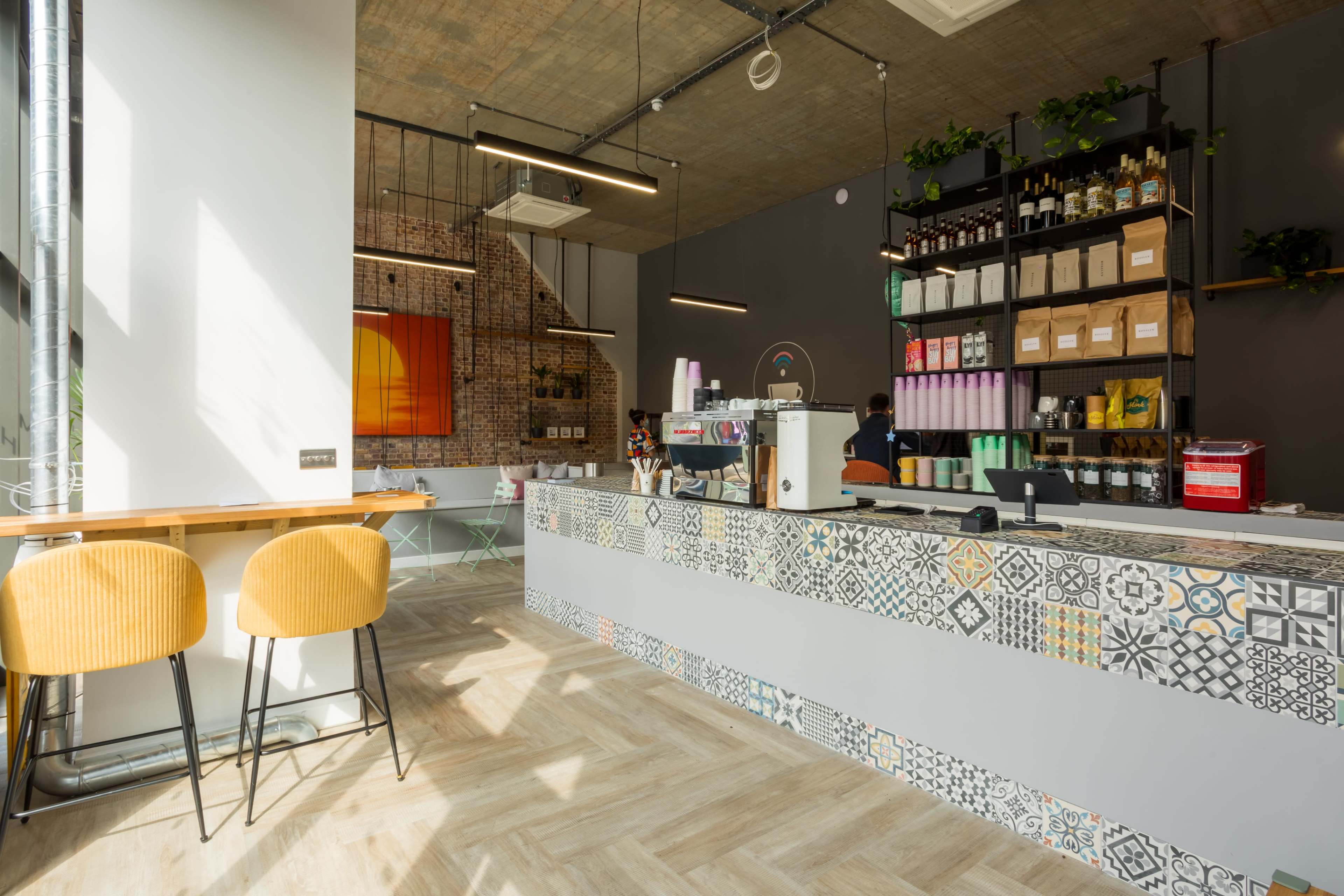 A modern café interior featuring a patterned counter, wooden seating, and shelves filled with various beverages and snacks.