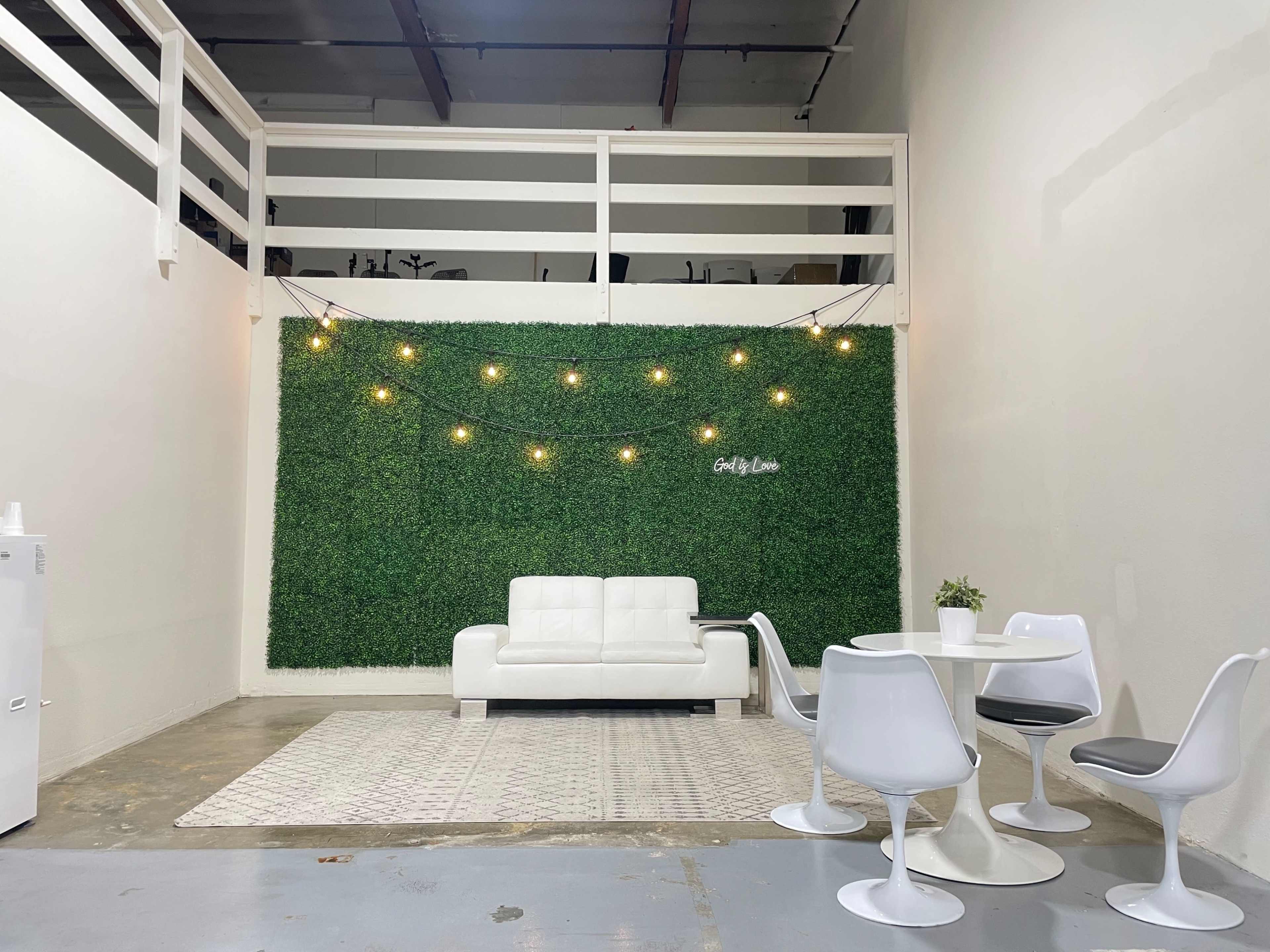 A modern indoor space with a white leather sofa, a round dining table, and two chairs, set against a green plant wall and decorative string lights.