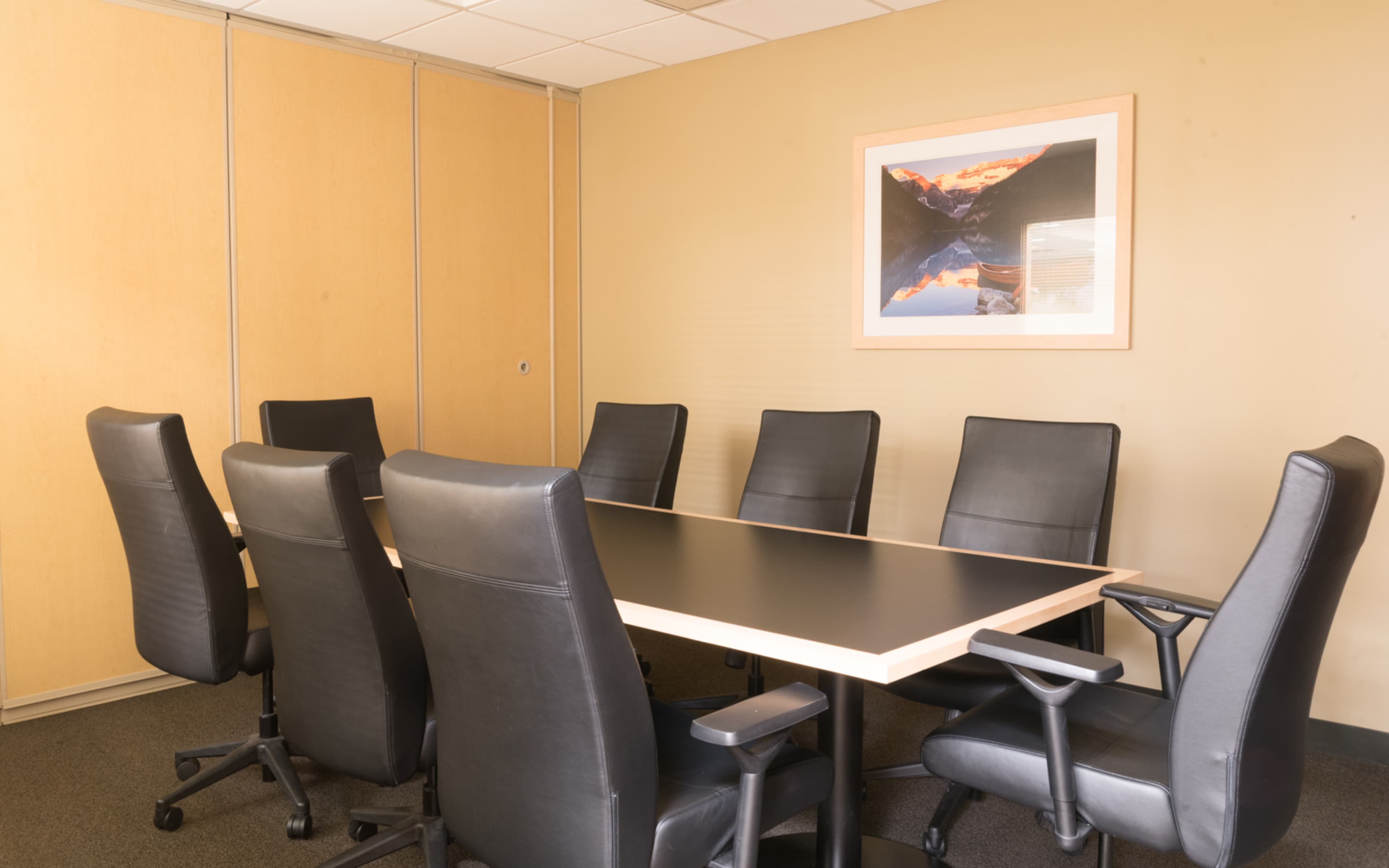 A conference room features a large black table surrounded by eight black chairs against a beige wall with a framed artwork hanging above.