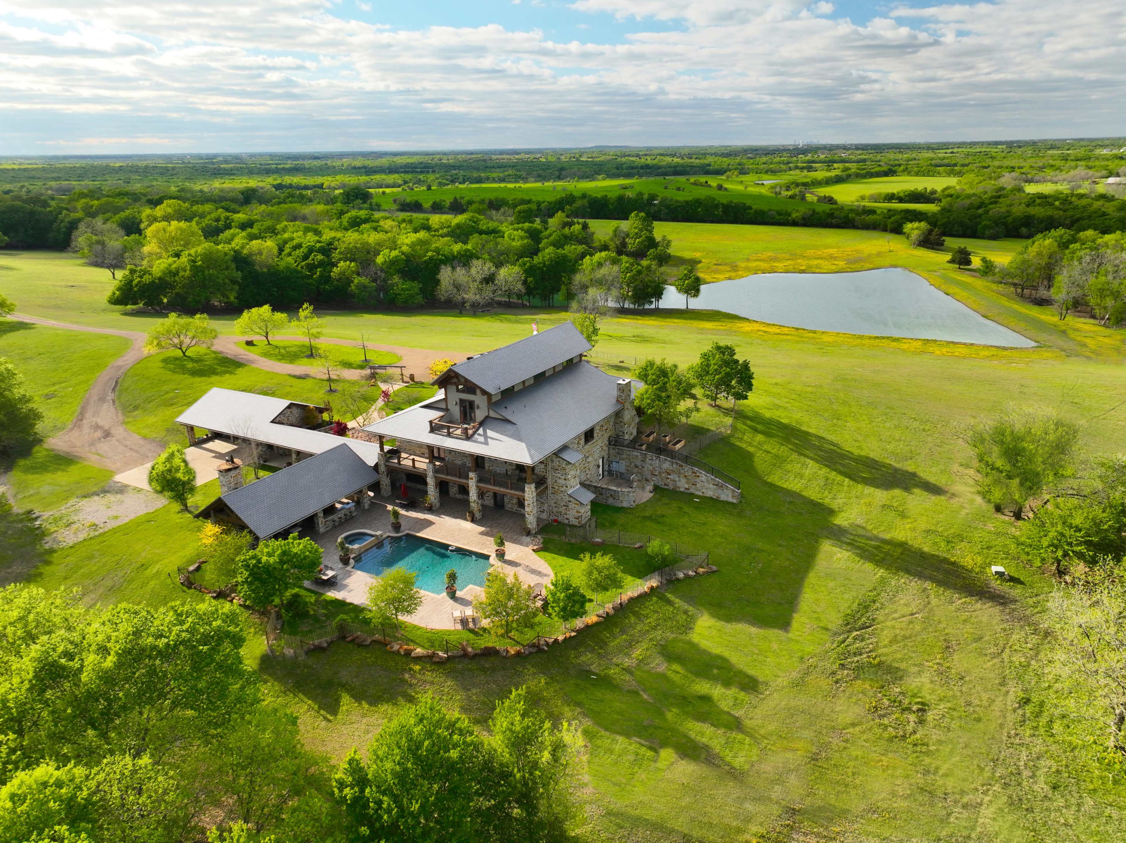 A large, modern house with a swimming pool, surrounded by greenery and a pond in a rural landscape.
