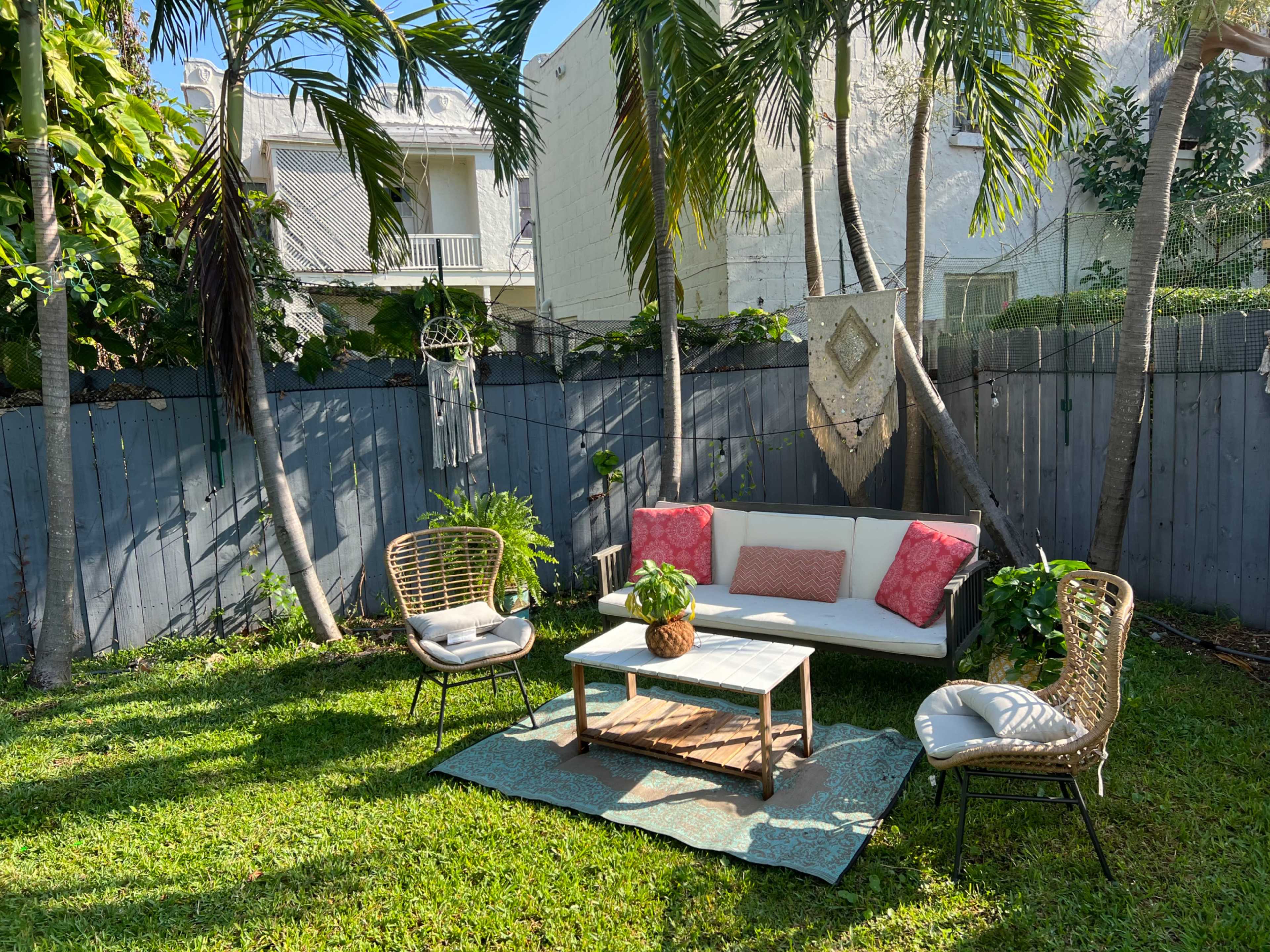 A small outdoor seating area features a white couch with pink cushions, a wooden coffee table, and three chairs surrounded by lush greenery and a wooden fence.