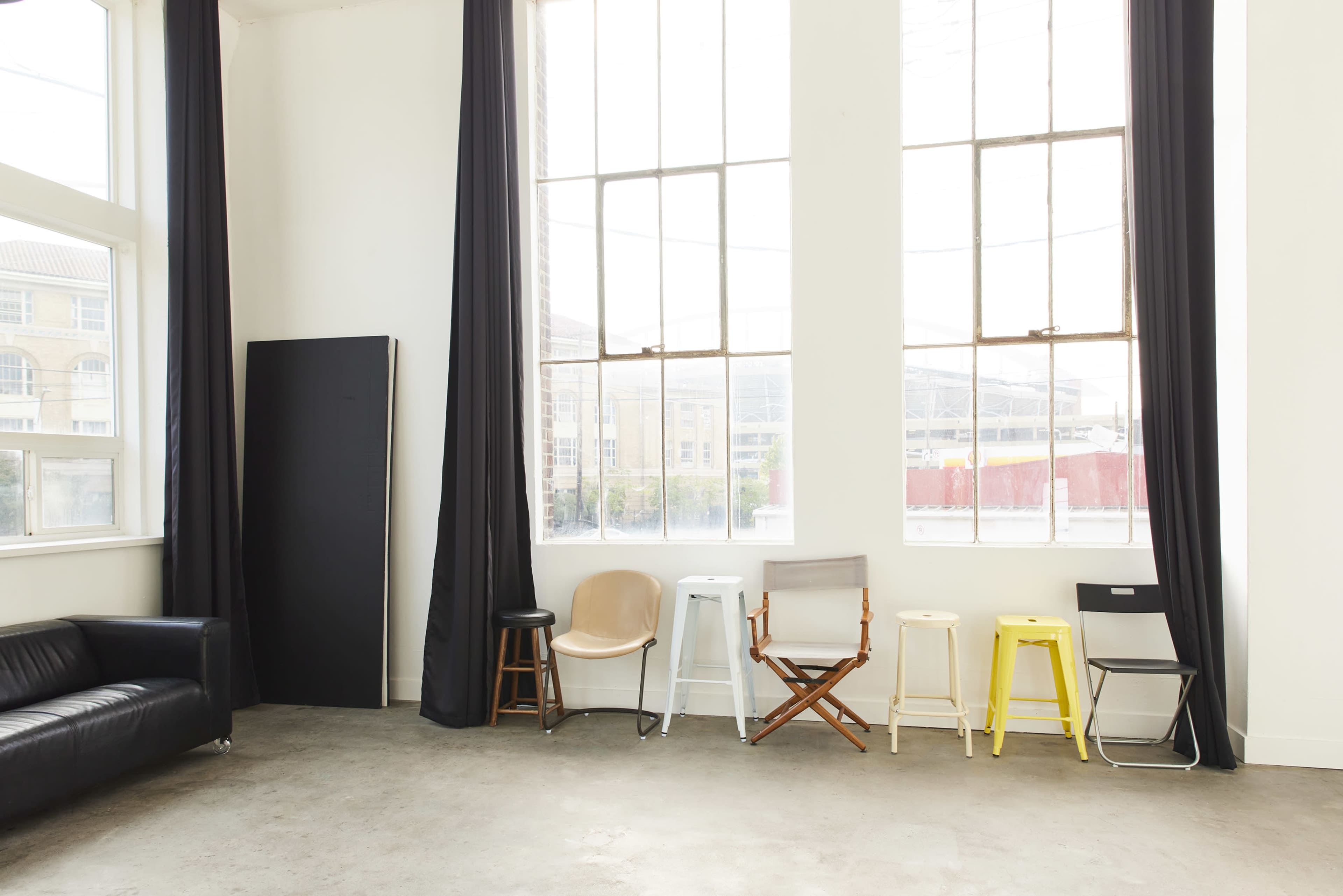 The image shows a bright room with large windows, featuring a black sofa and a variety of chairs lined up against the wall.
