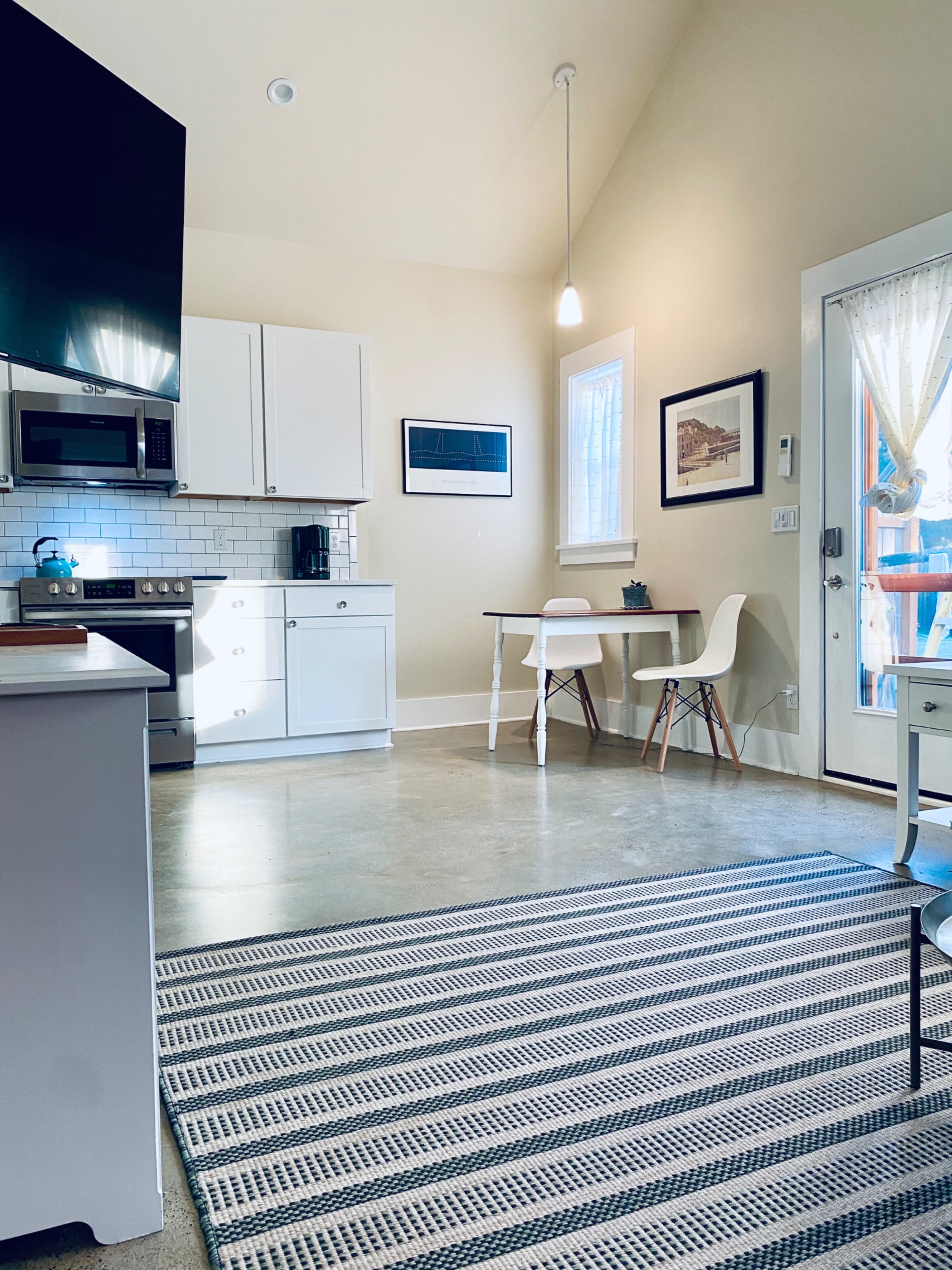 The image shows a modern kitchen with white cabinets, a stainless steel stove, and a small dining table beside a door leading outside.