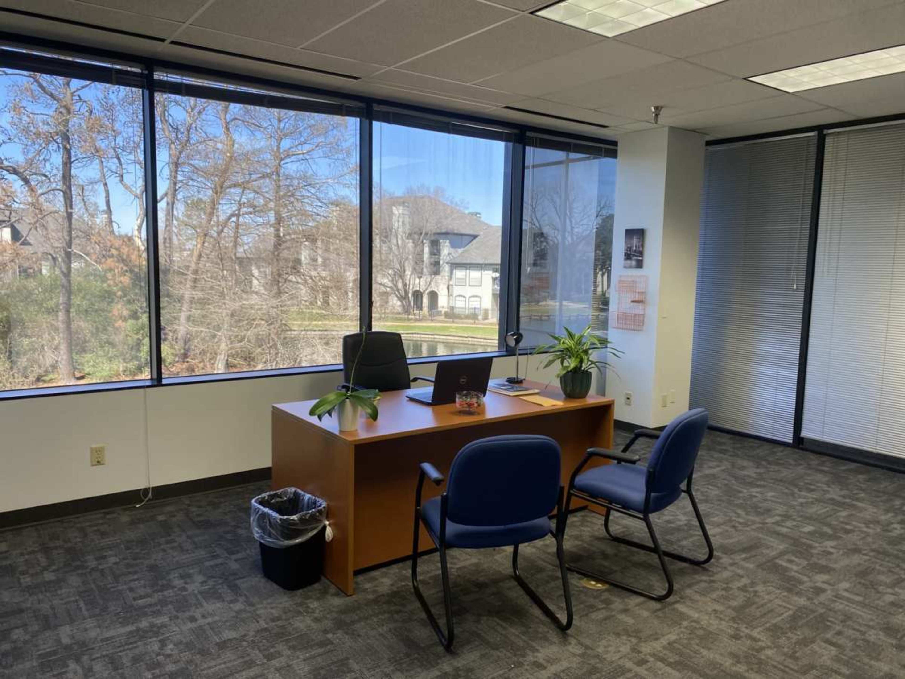 The image shows a small office space with a wooden desk, two blue chairs, and large windows overlooking a green area outside.