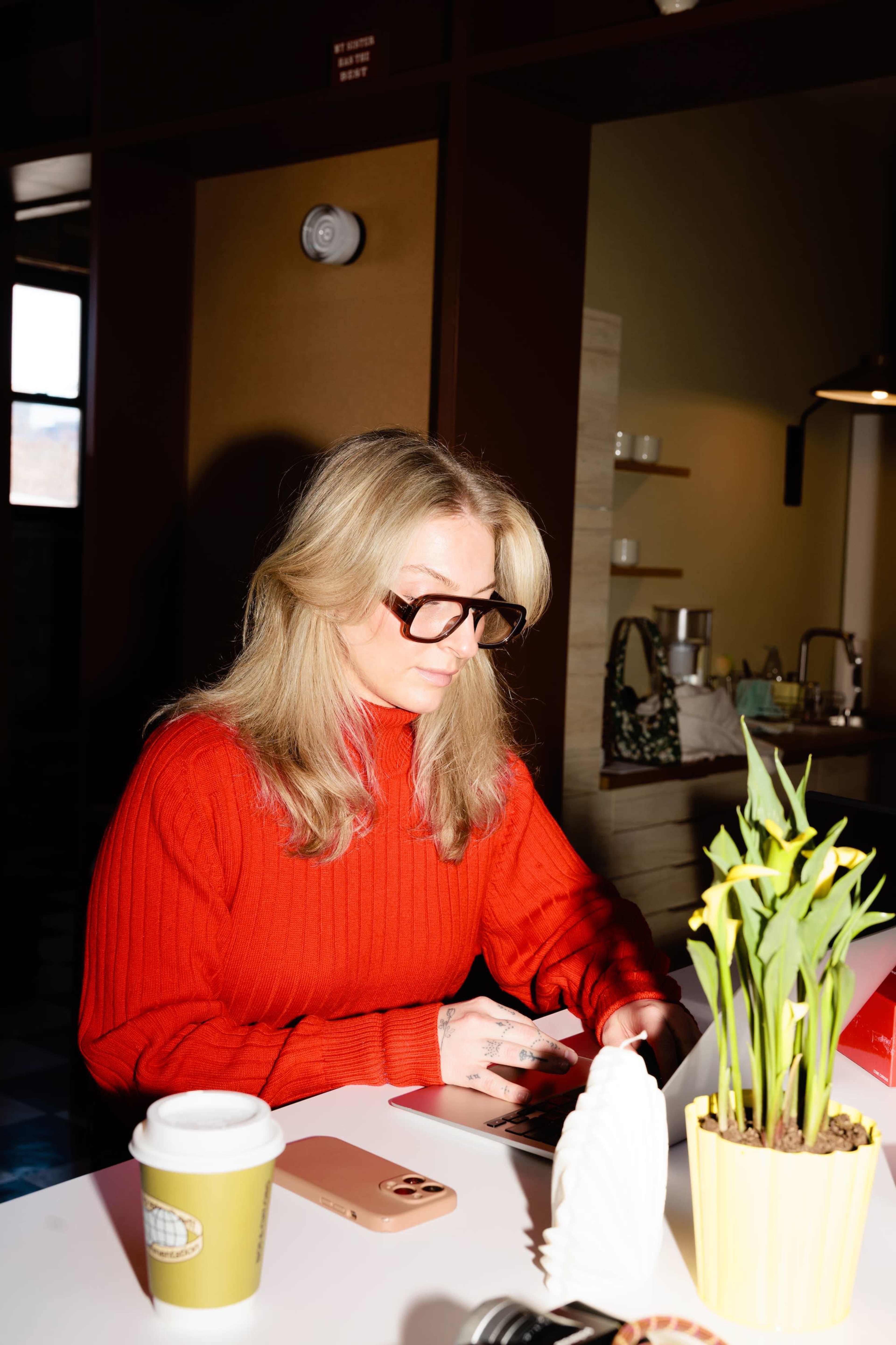 A woman with long blonde hair sits at a table, wearing a red sweater and large glasses, while using a smartphone next to a potted plant and a coffee cup.