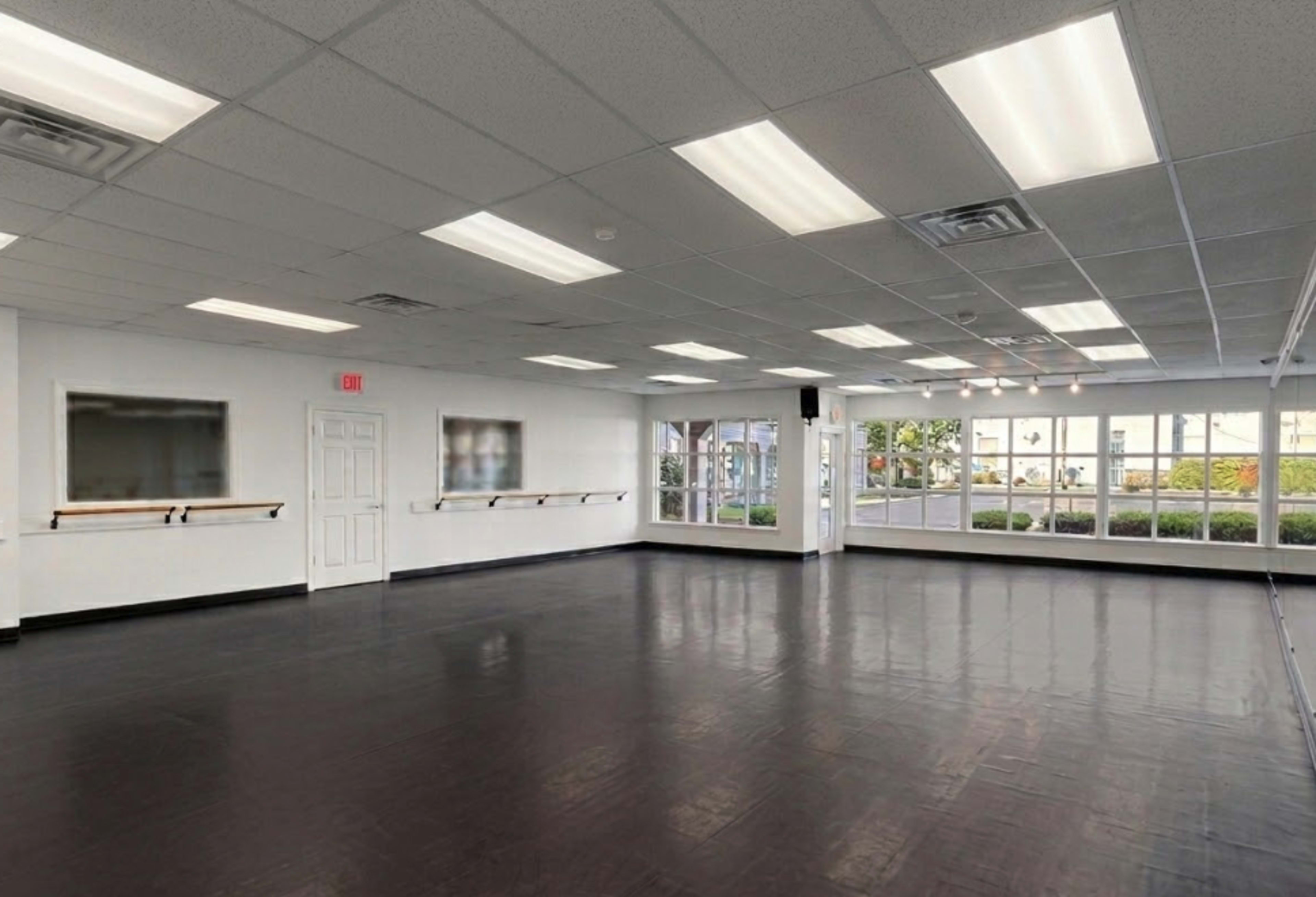 The image shows a spacious dance studio with a polished black floor, mirrored walls, and large windows letting in natural light.