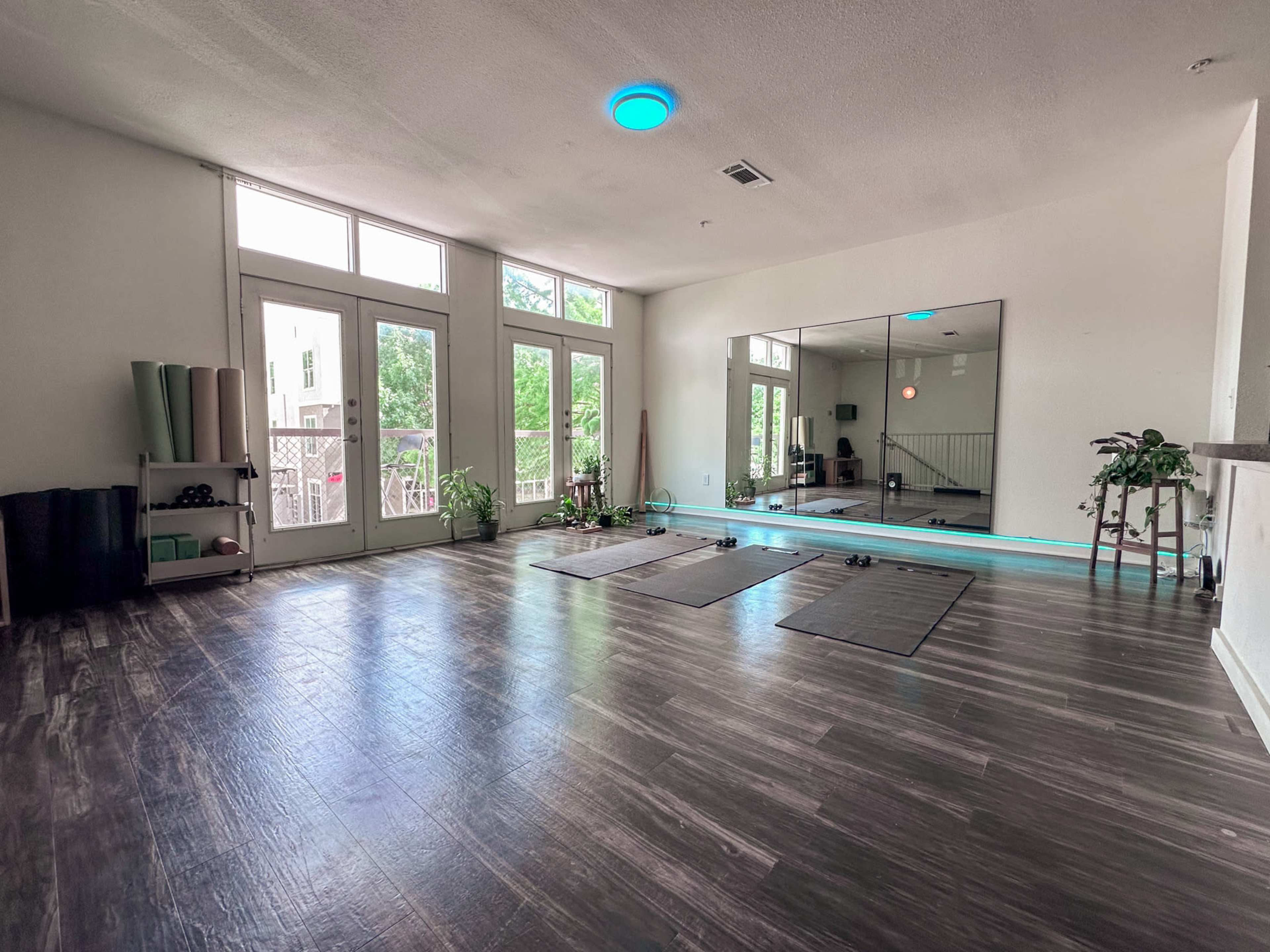 The image shows a spacious, well-lit room with hardwood floors, two yoga mats on the ground, and large windows that provide natural light.