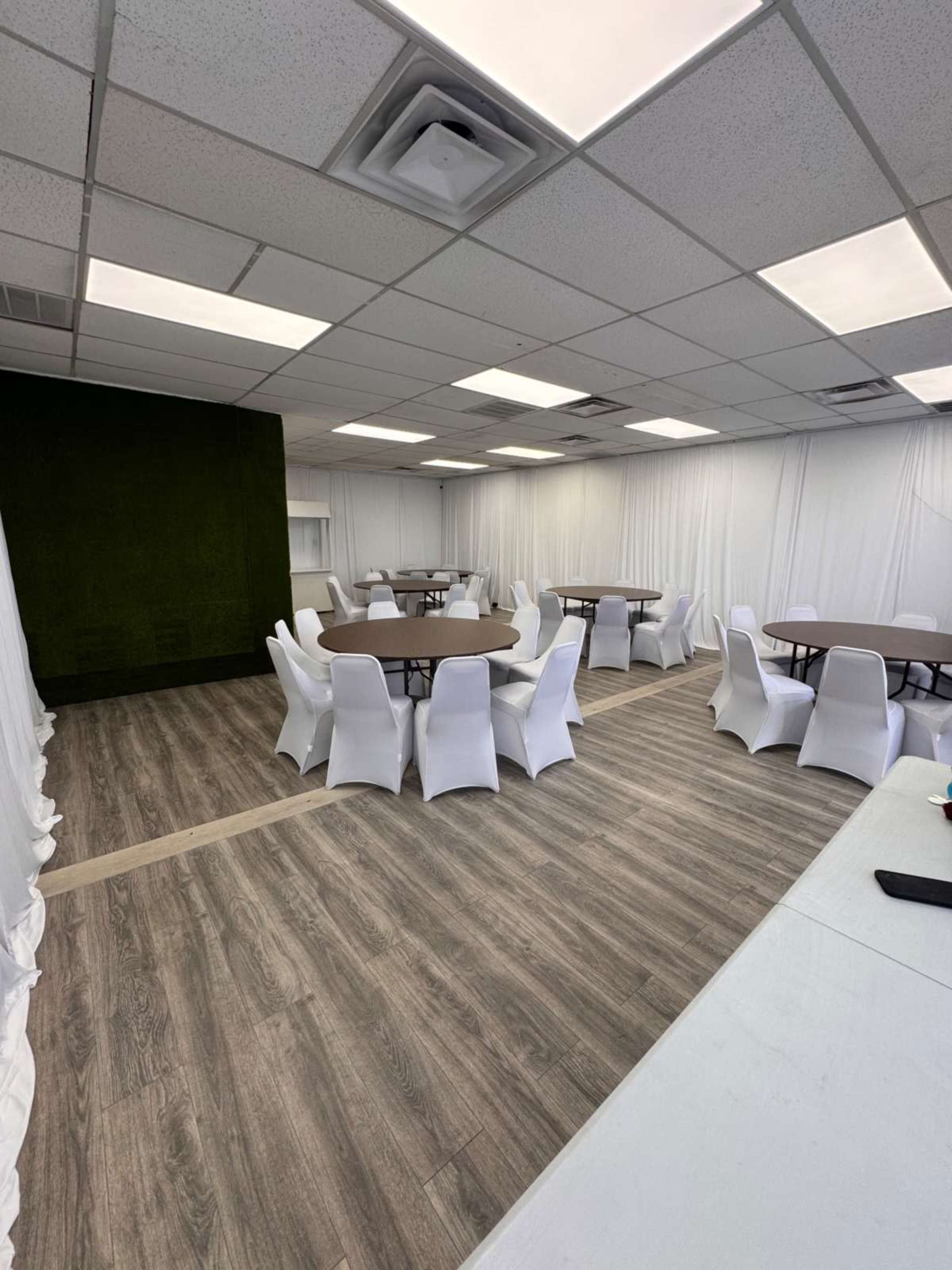 The image shows a spacious event room featuring multiple round tables surrounded by white chairs, with a textured green wall in the background.