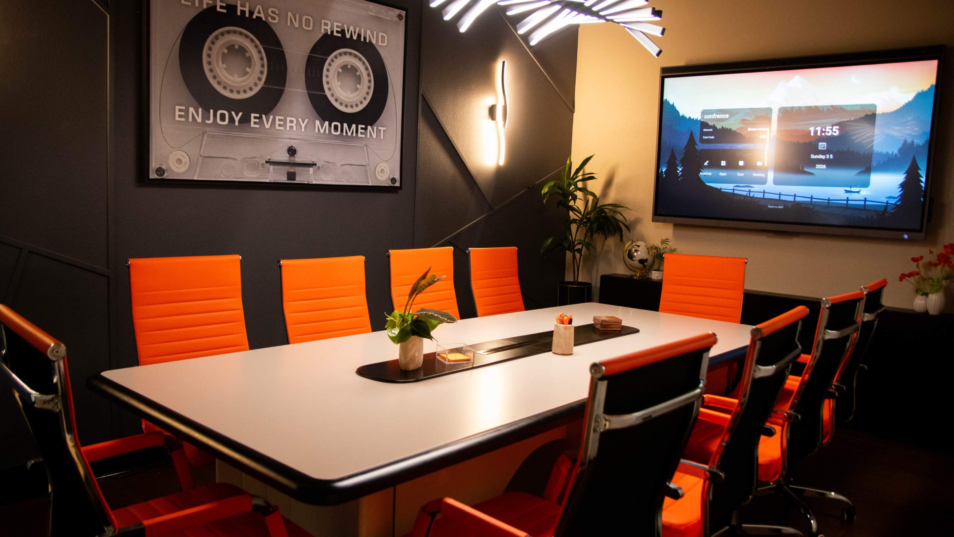 The image shows a modern conference room with a large table surrounded by orange chairs, featuring a digital display and decorative wall art.