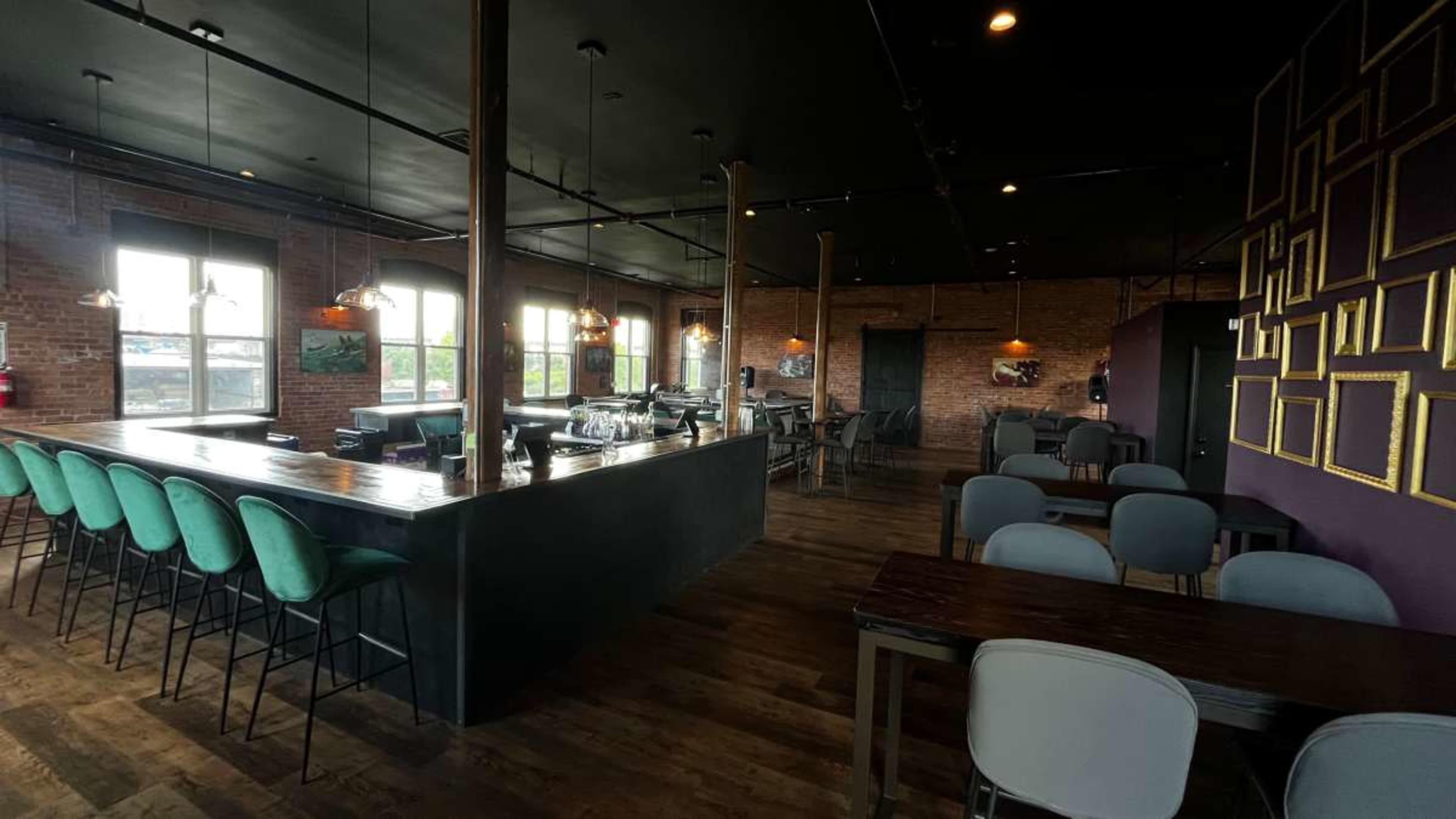 A spacious, modern bar area with wooden floors, black walls, and tall stools lined up along a bar counter, illuminated by pendant lights.