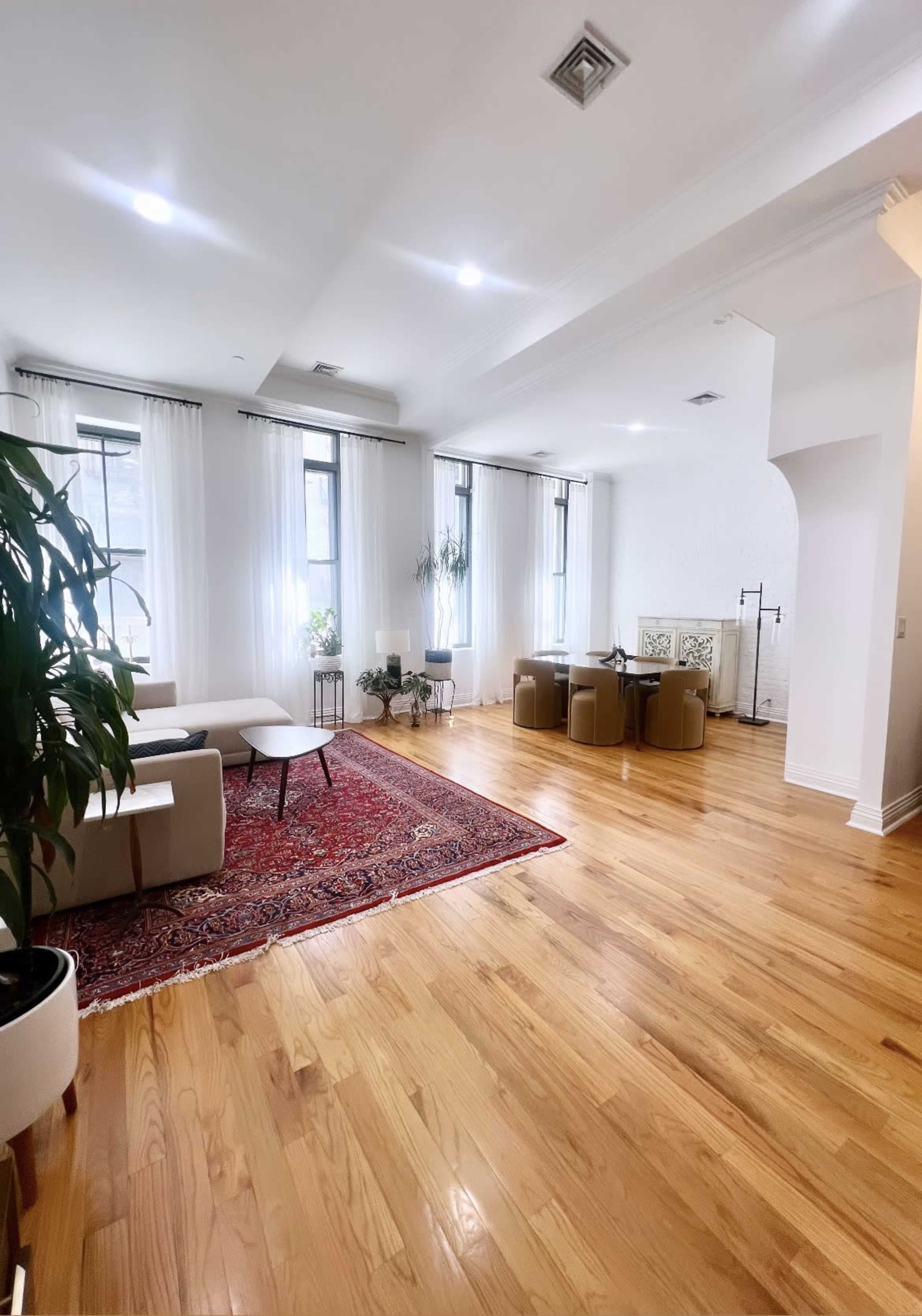 A spacious living area with wooden flooring, a red area rug, two seating areas, large windows with sheer curtains, and indoor plants.