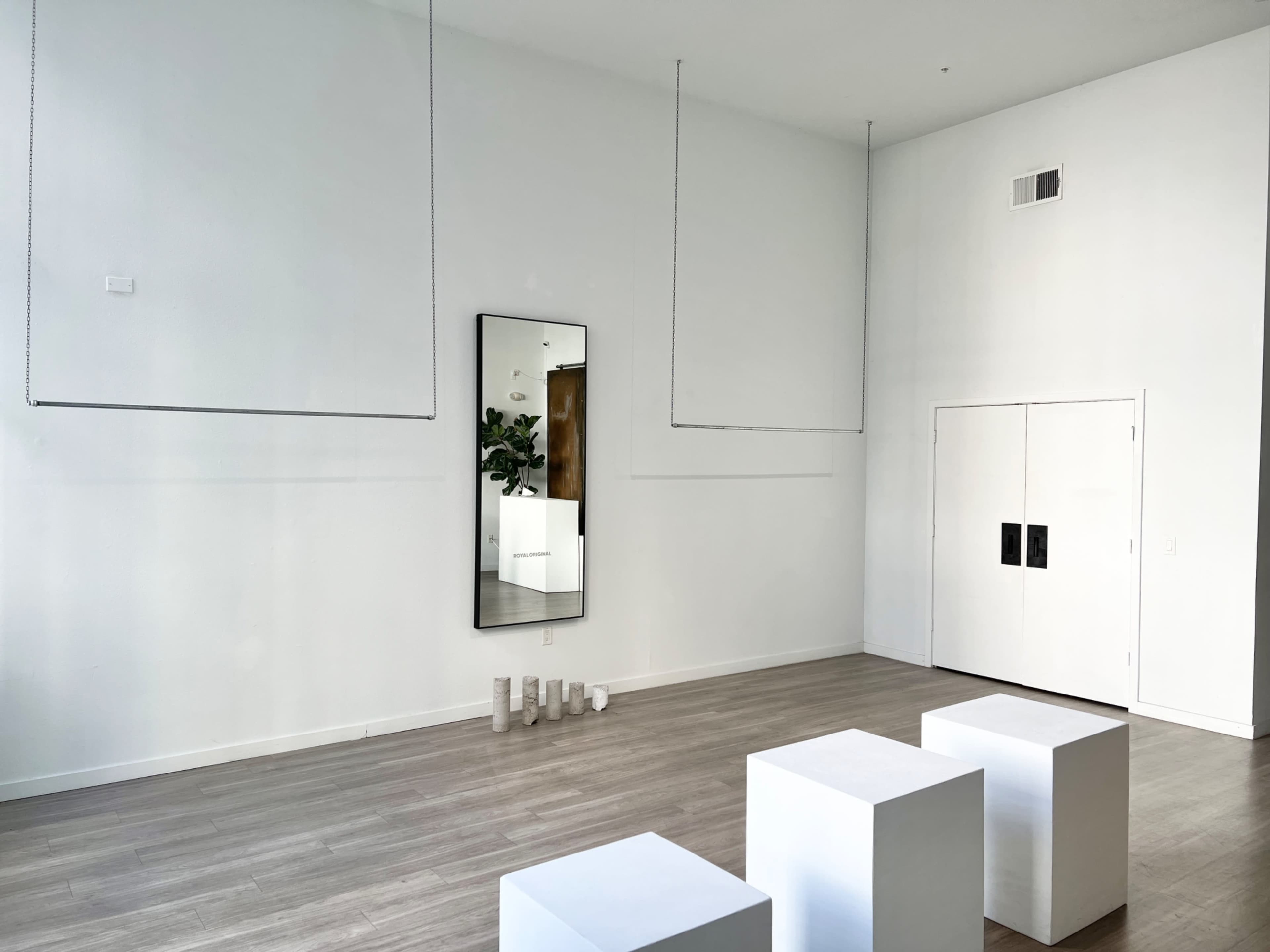 A minimalist interior space with white walls, hanging elements, a large mirror reflecting the room, three white cubes, and a closed double door.