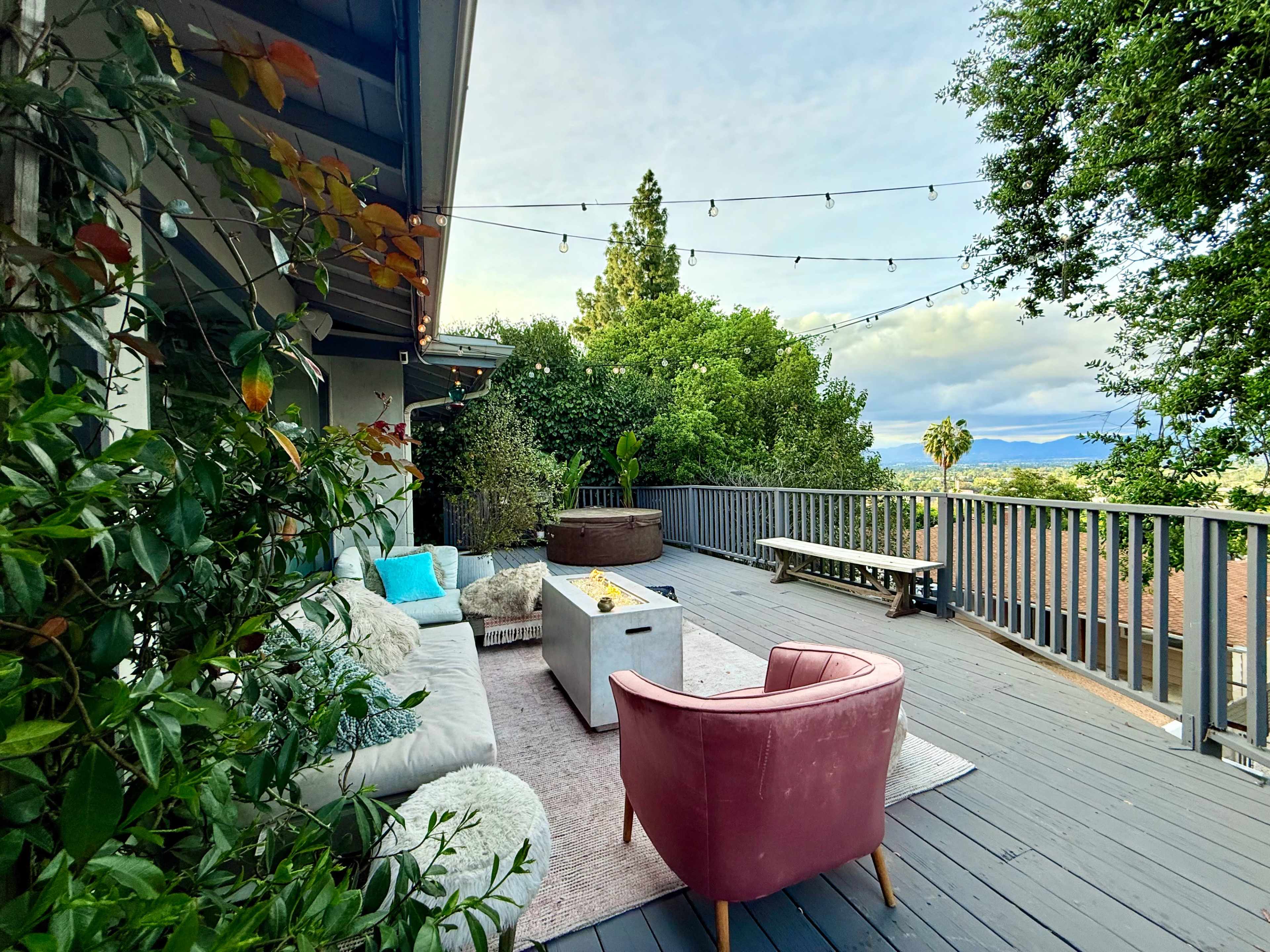A wooden deck with a cozy seating area, potted plants, and string lights offers a view of trees and distant hills.