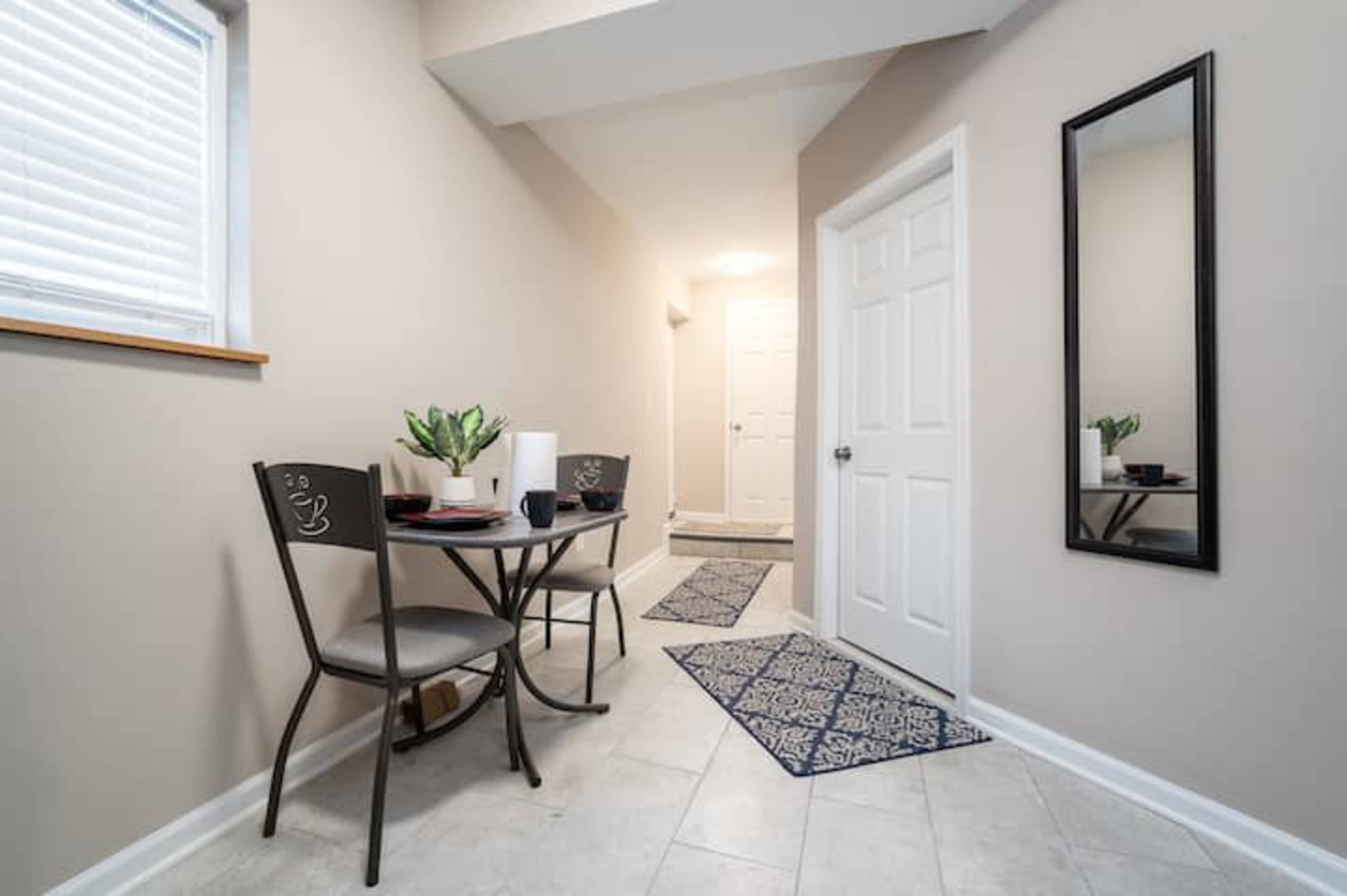 A small hallway features a table and two chairs alongside a mirror, with a door leading to another room at the end.