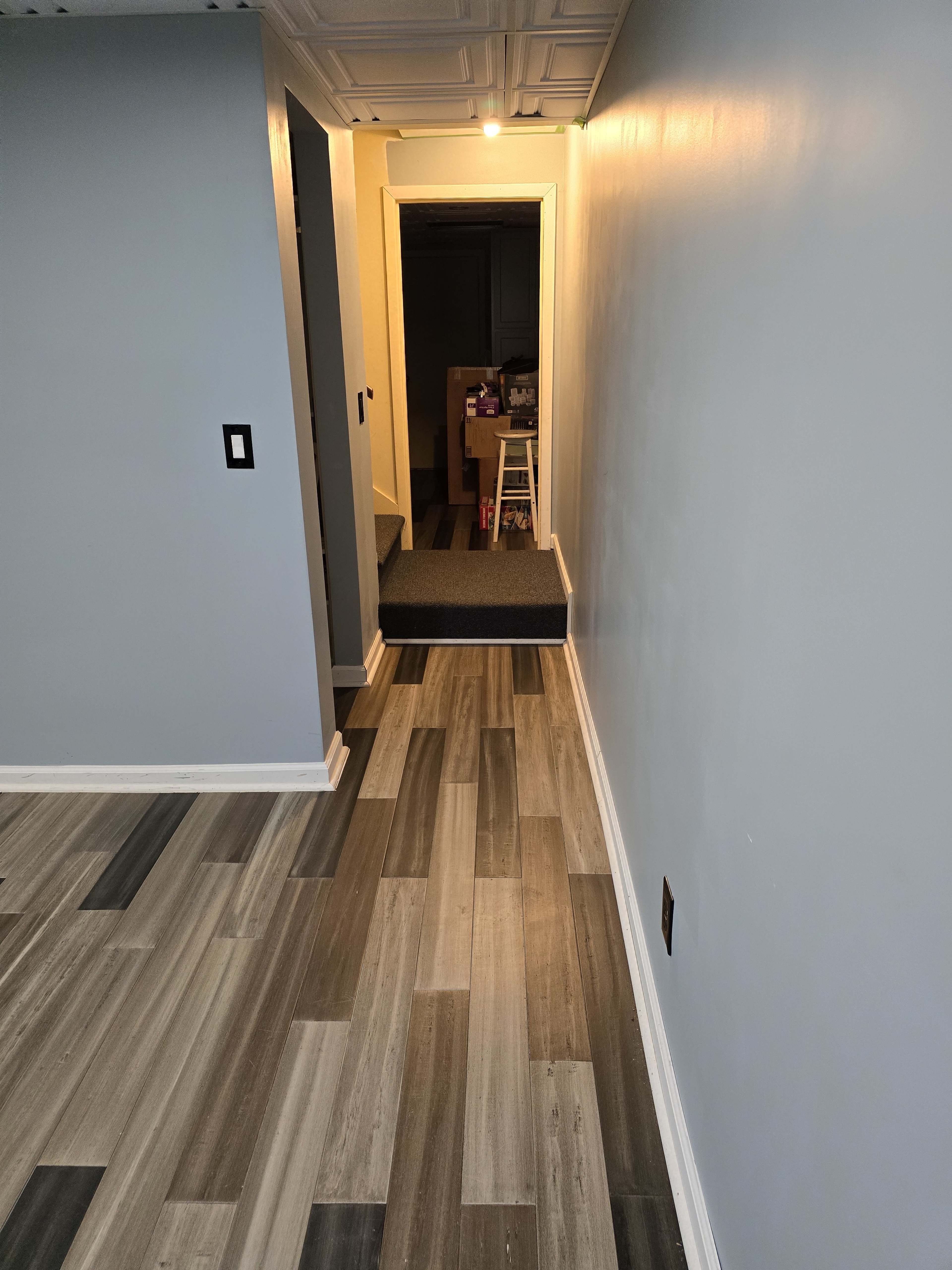 A hallway with light gray walls features a staircase leading to a darker area and a floor of alternating gray plank tiles.