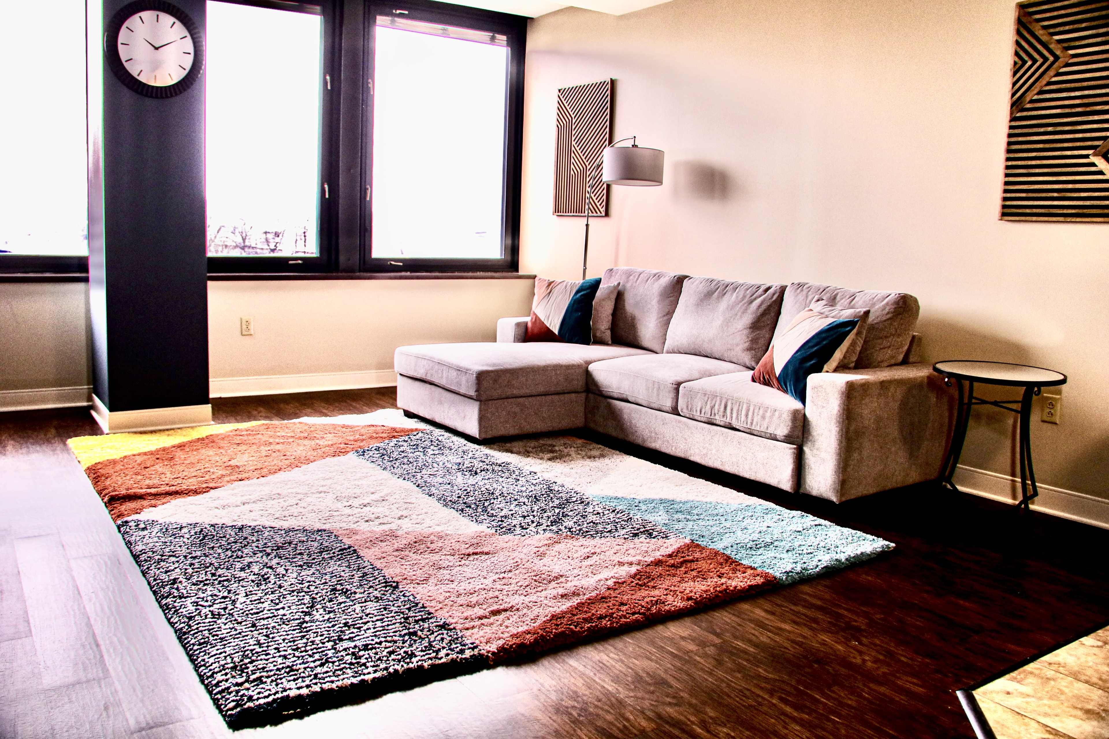 A modern living room features a sectional sofa, a large patterned rug, and a floor lamp next to a window with city views.