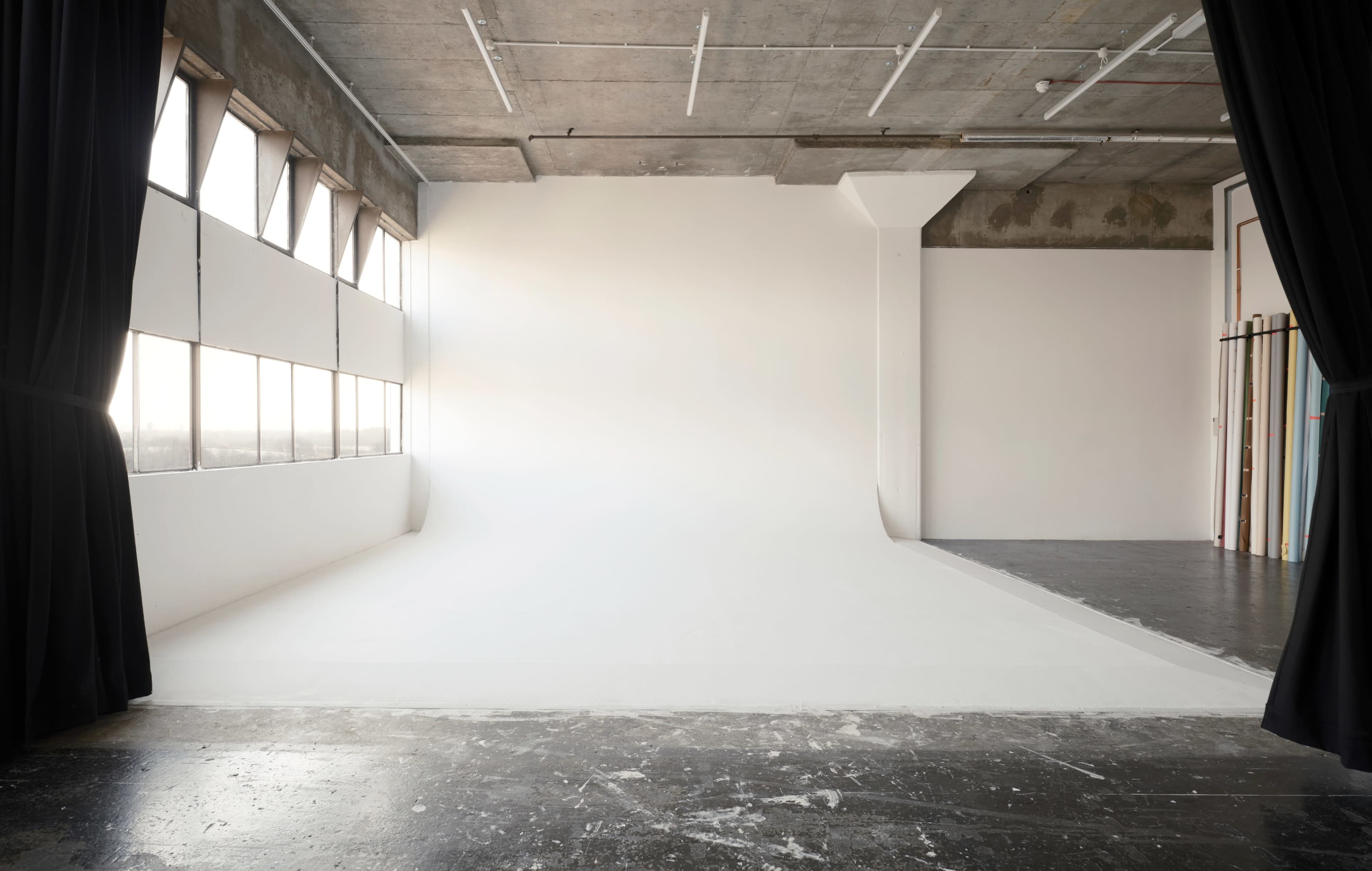 A spacious studio with large windows, a seamless white backdrop, and a concrete ceiling.