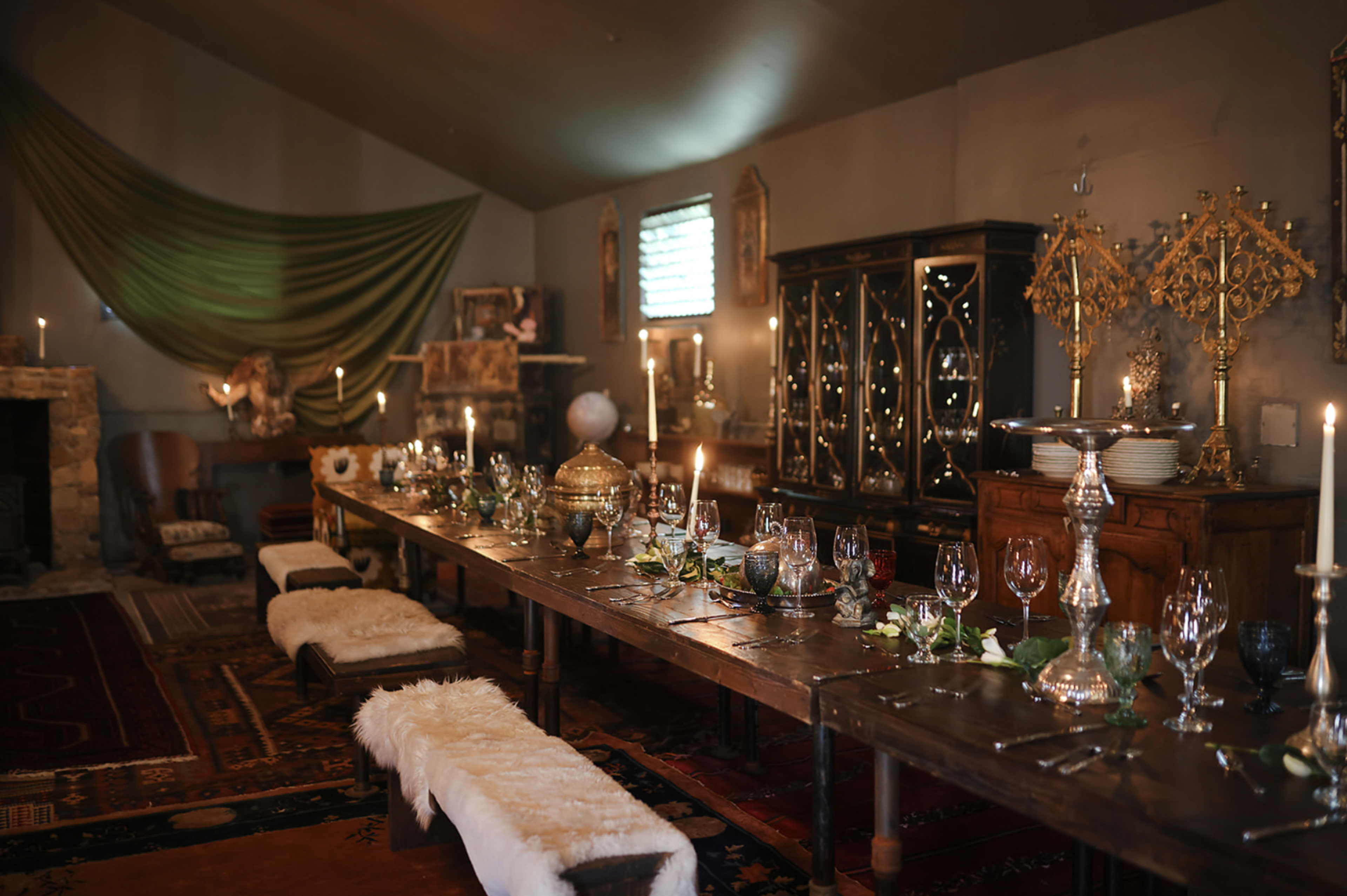 A long wooden dining table, set with elegant glassware and candlelight, is positioned in a dimly lit room adorned with decorative furnishings and textiles.