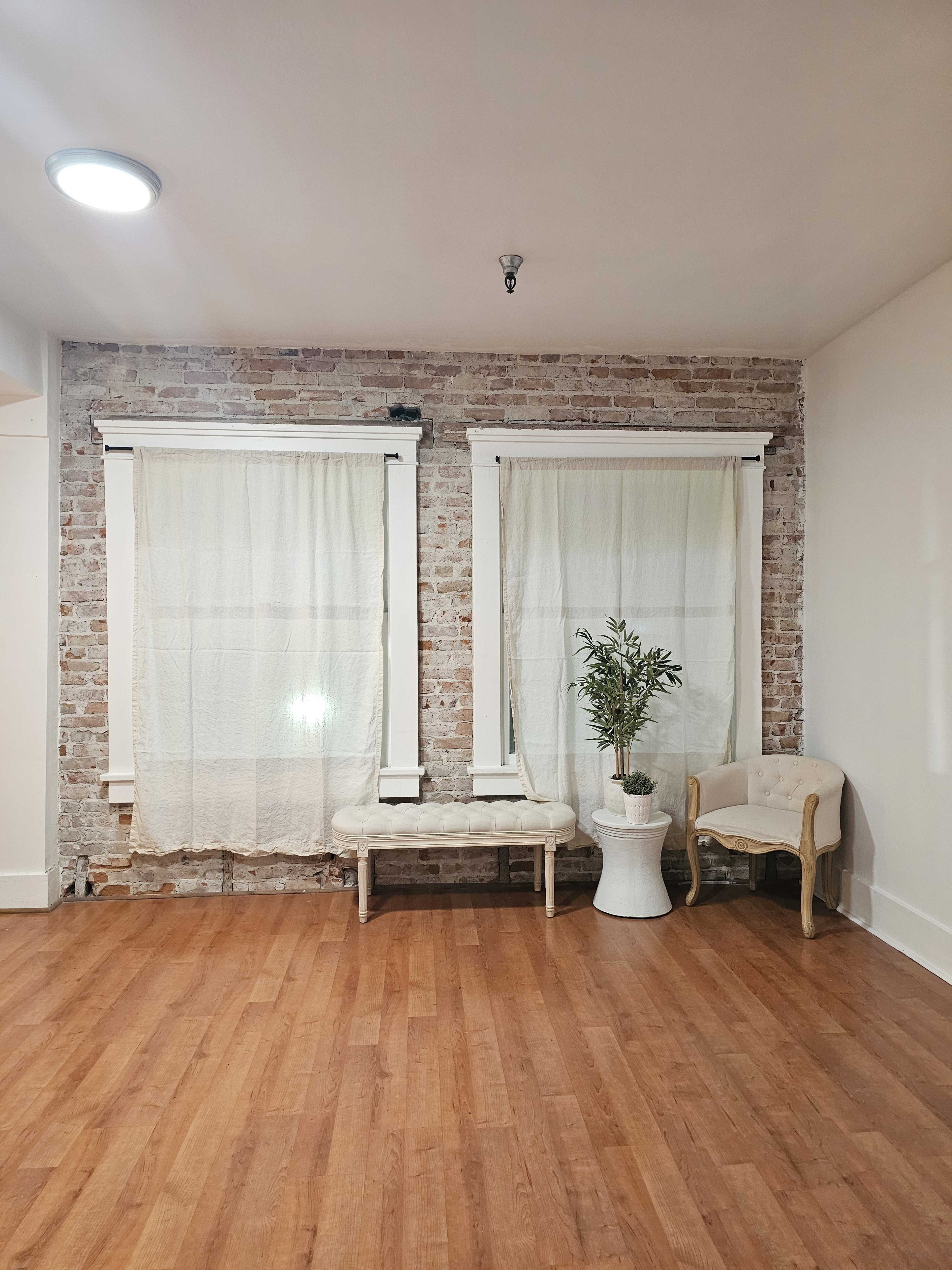 A spacious room features two large windows with light curtains, a brick wall, a wooden floor, a white plant pot with a plant, and a small bench.