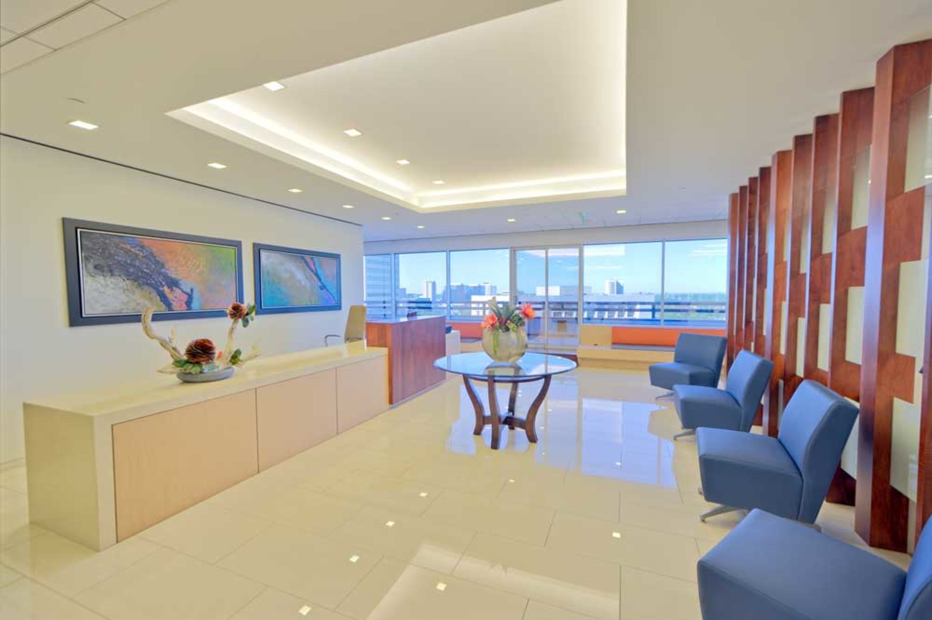 The image shows a modern office reception area with a glass-top table, blue chairs, and large windows offering a view of the city skyline.