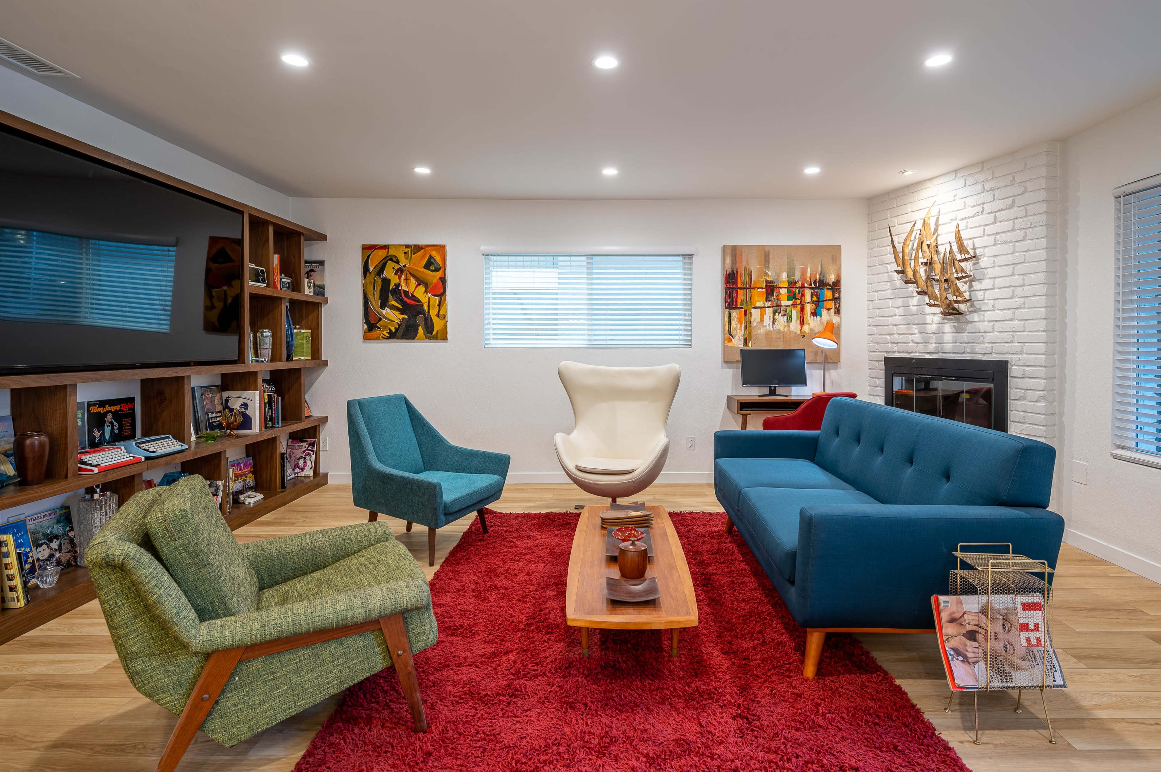 A modern living room featuring a mixture of teal and green seating, a wooden coffee table, a red rug, and shelves with decorative items against a white brick wall.